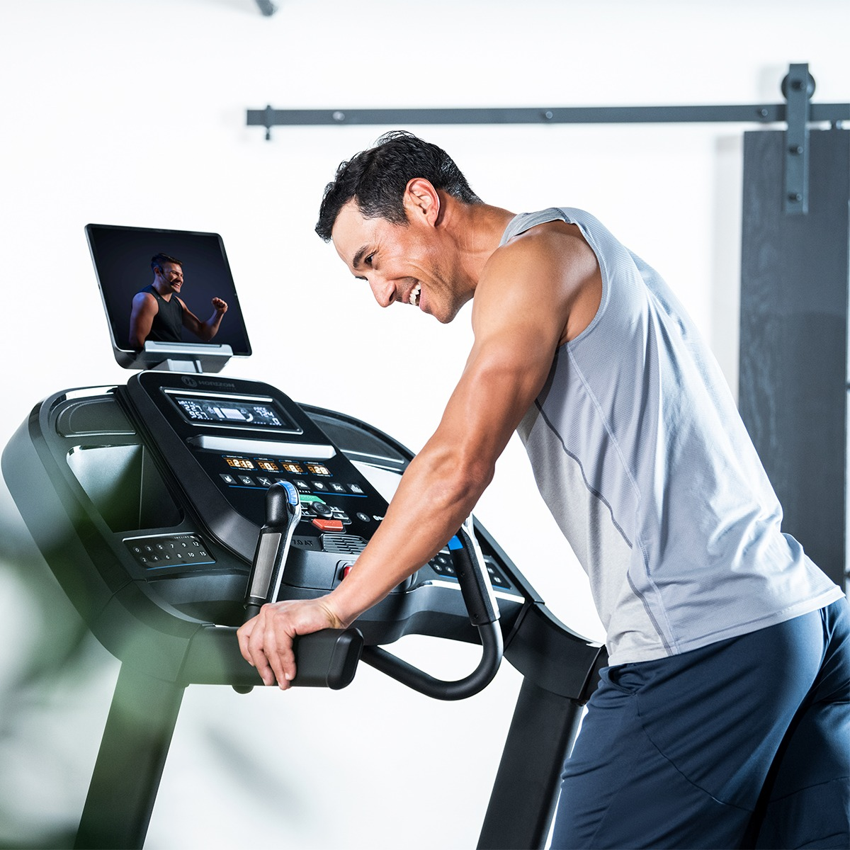 Mężczyzna na bieżni, patrzy na ekran. Uśmiechnięty. Szara koszulka, ciemne szorty. Siłownia, biała ściana, drewniane drzwi.