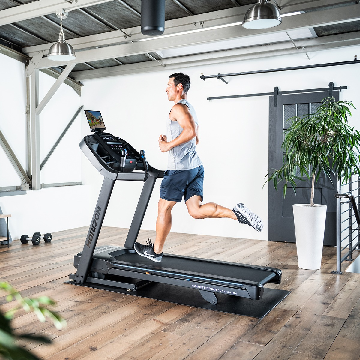 Mężczyzna biegnący na bieżni w siłowni, ubrany w odzież sportową. Białe i drewniane wnętrze.