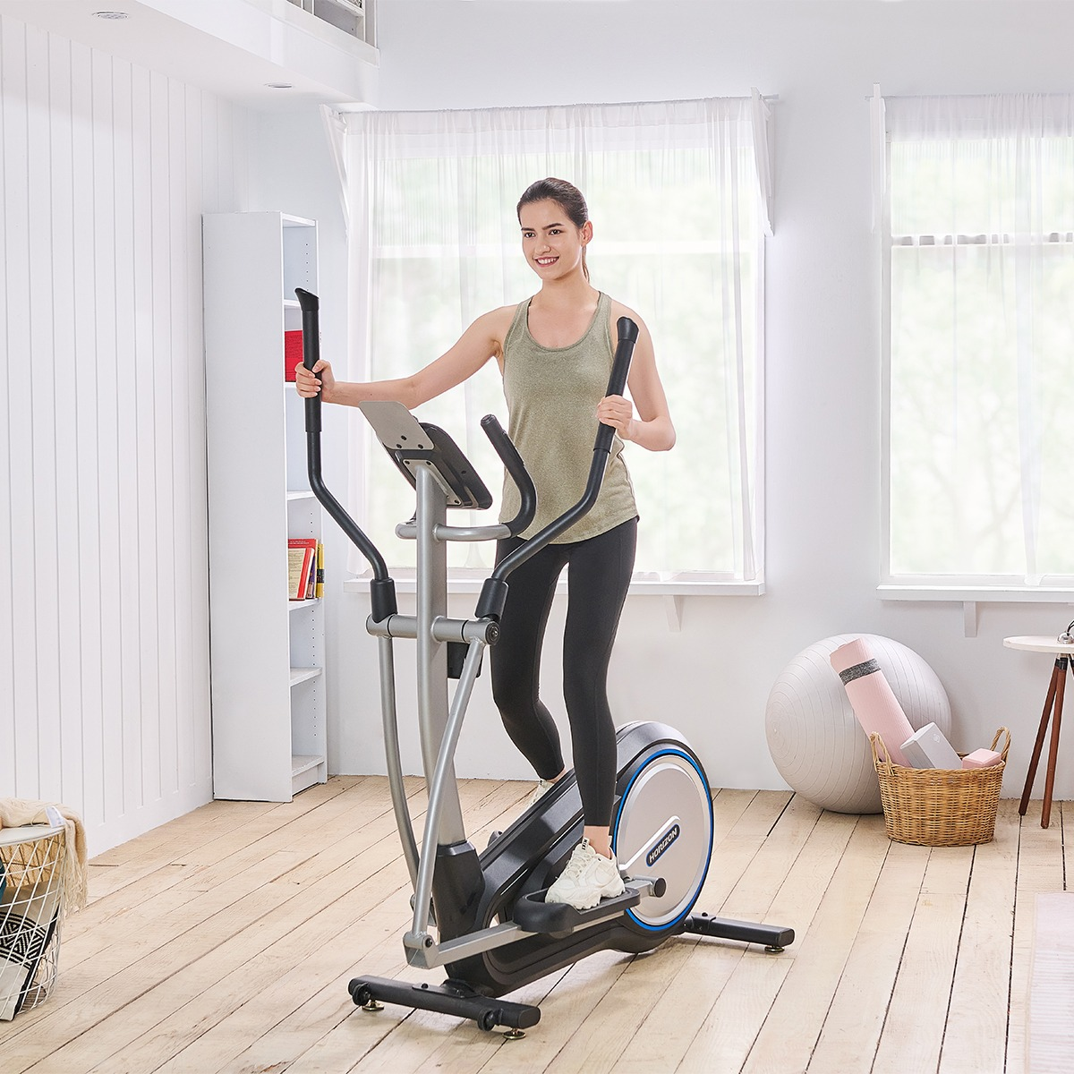 Kobieta ćwiczy na orbitreku. Pokój jest jasny, z dużymi oknami i drewnianymi podłogami.