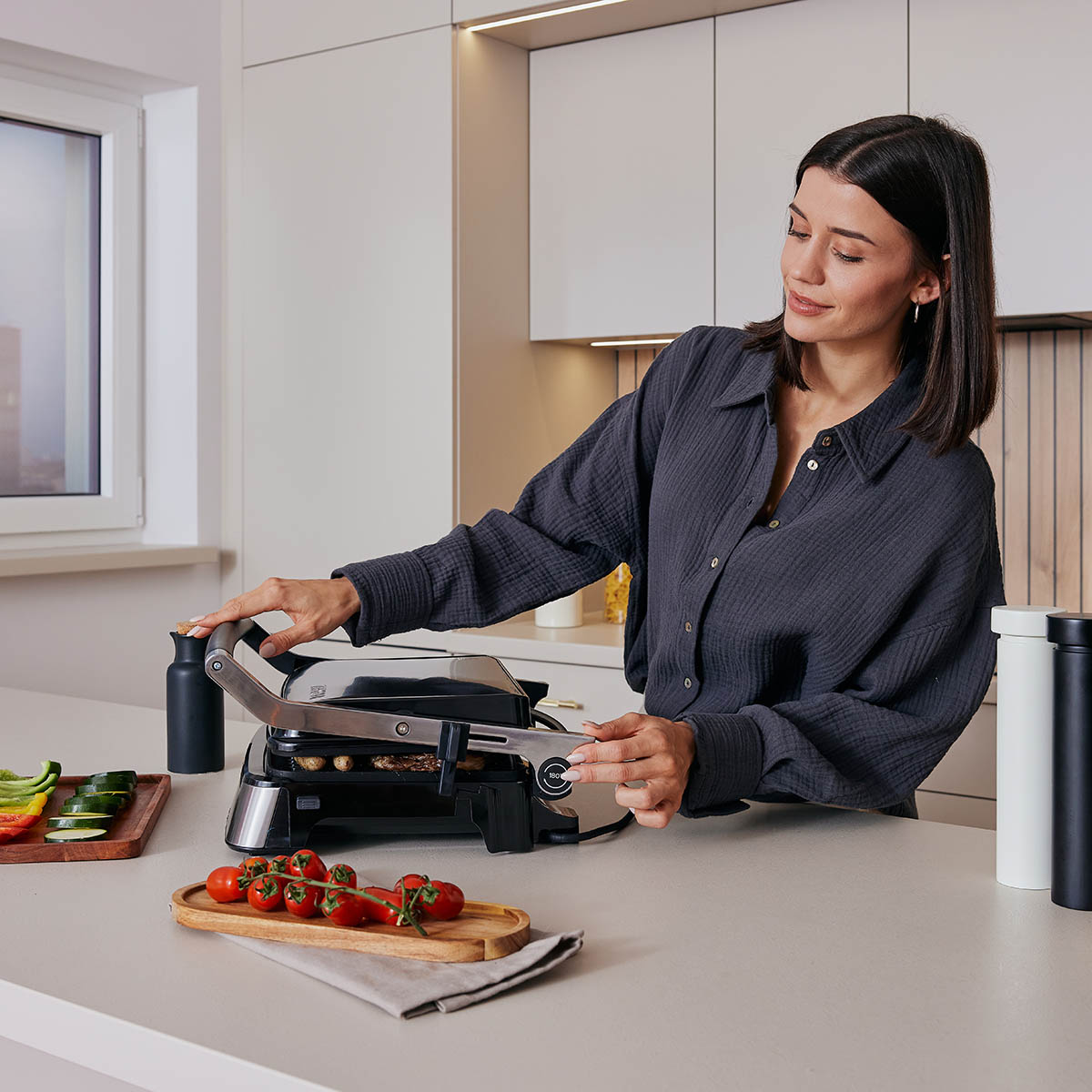 Frau grillt in einer Küche. Sie benutzt einen Grill, mit Gemüse und einer Flasche auf der Theke.
