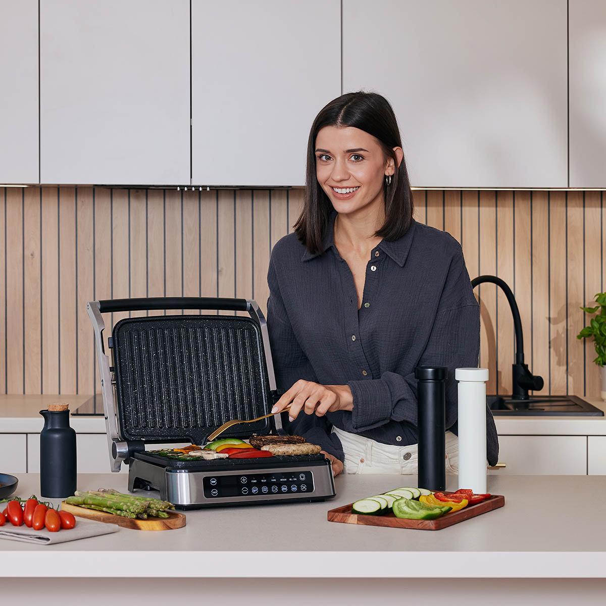 Frau grillt Gemüse auf einem Indoor-Grill. Küchentheke mit Essen und Utensilien, lächelt in die Kamera.