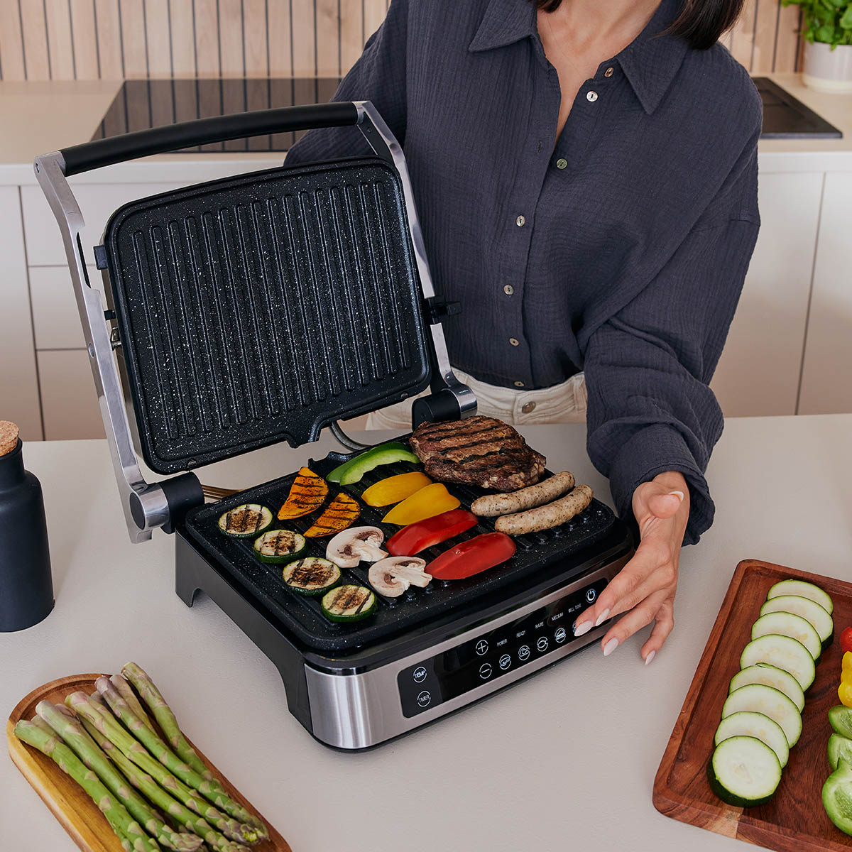 Frau grillt Gemüse, Würstchen und Fleisch auf einem offenen Grill. Verschiedene Beilagen sind angeordnet.