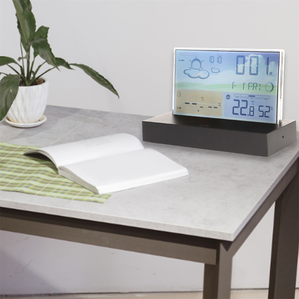 Ein Schreibtisch mit Wetterstation, offenem Buch und Pflanze im Hintergrund.