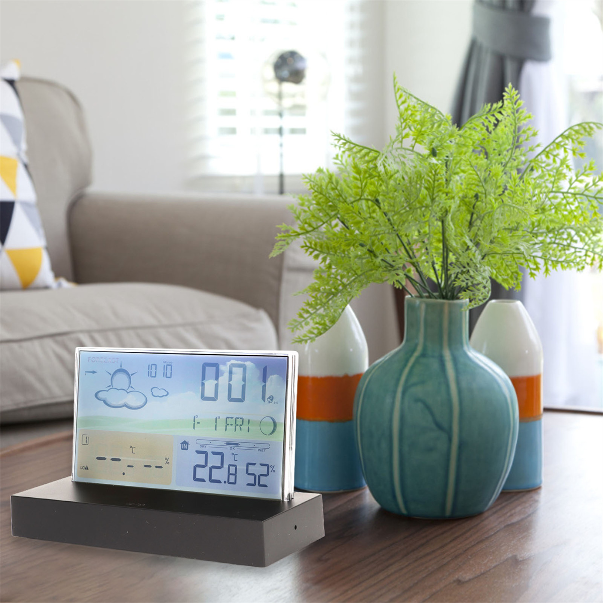 Eine digitale Wetterstation auf einem Tisch, mit einer Vase und grünen Pflanzen.