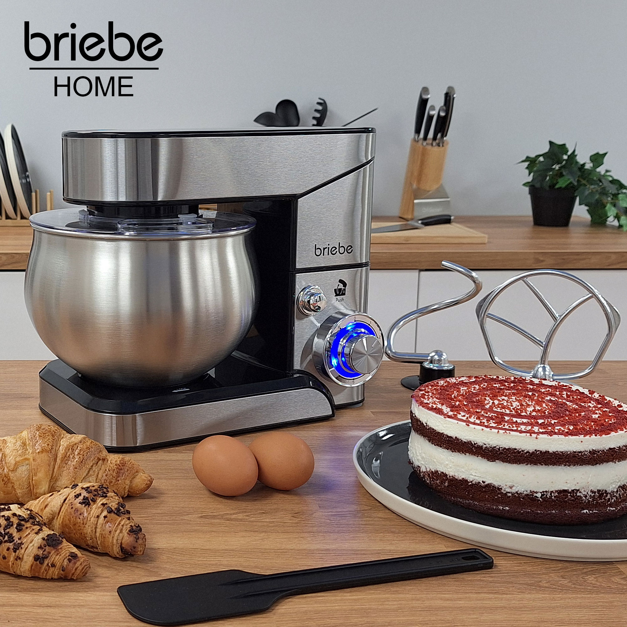 Ein Edelstahlmixer mit Kuchen, Croissants, Eiern und einem Spatel auf einem Holztisch.