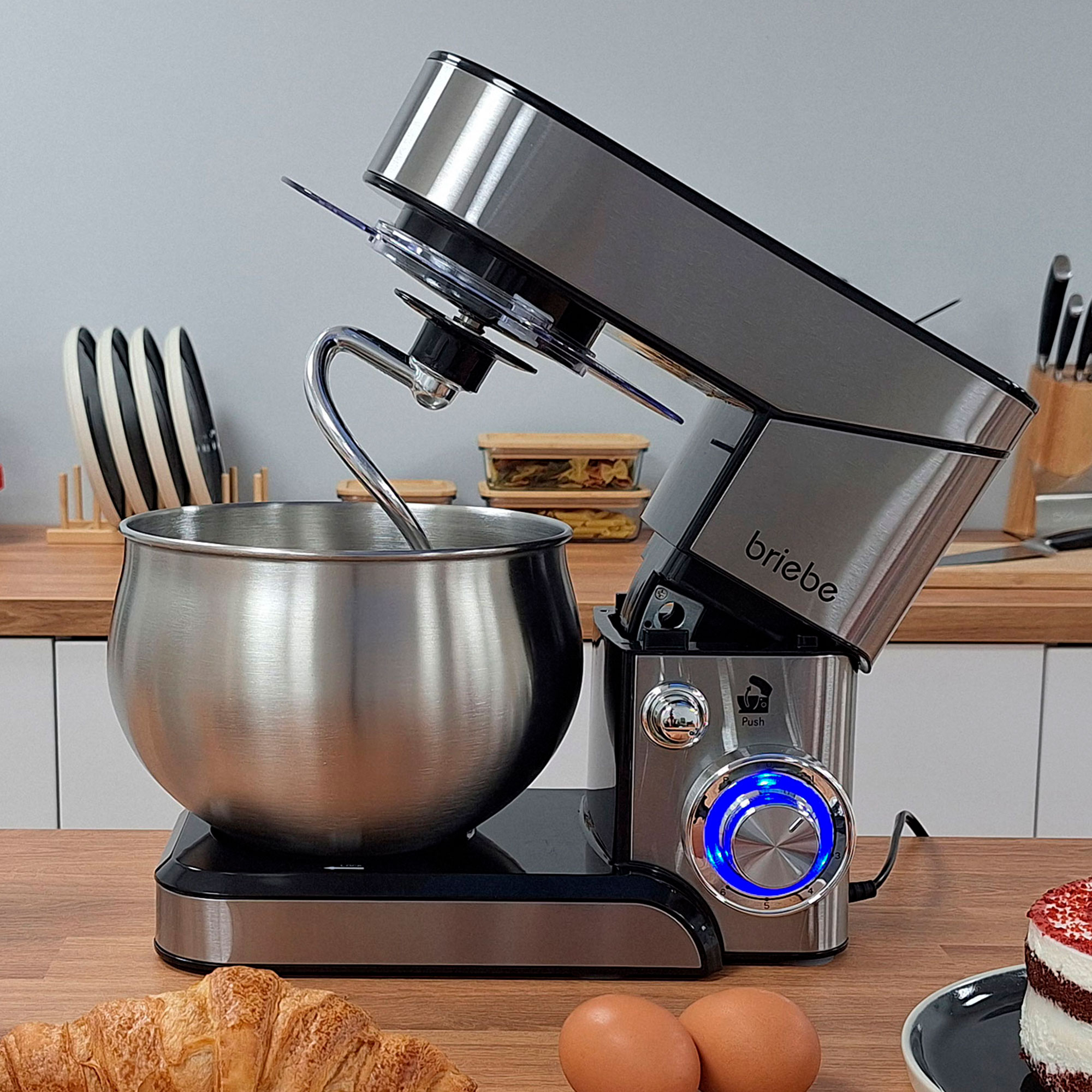 Edelstahl-Küchenmaschine mit Schüssel, Haken und blauem Licht. Croissant und Eier auf Holzoberfläche.