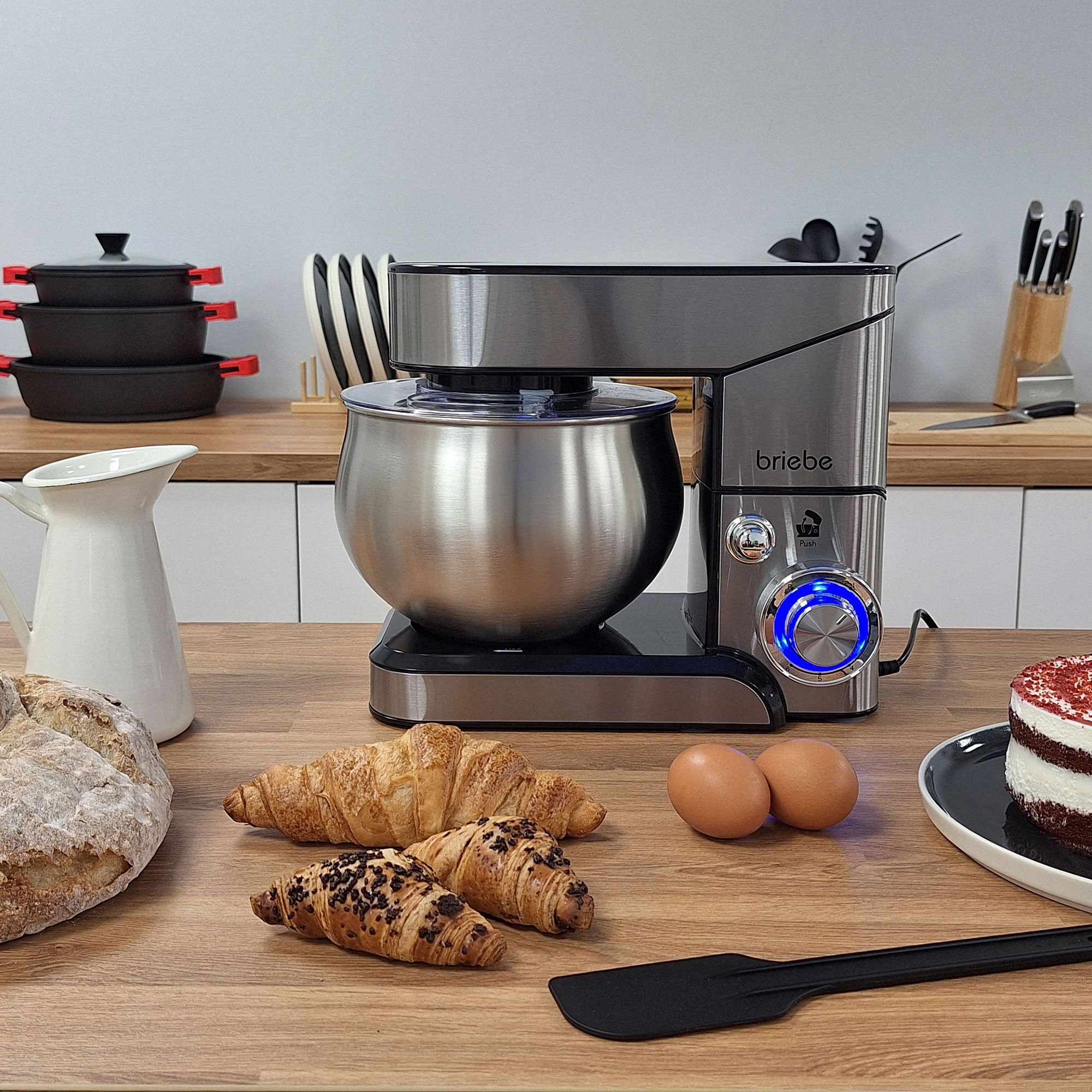 Ein Küchenmixer mit Edelstahlschüssel und Zubehör. Zutaten befinden sich auf der hölzernen Arbeitsplatte.