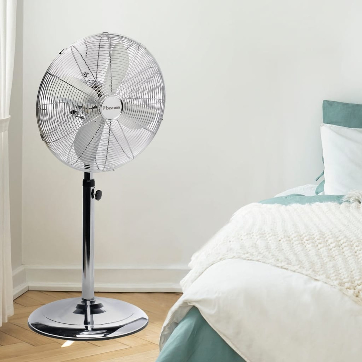 Un ventilatore a colonna cromato in una camera da letto. Un letto con una coperta bianca sullo sfondo.