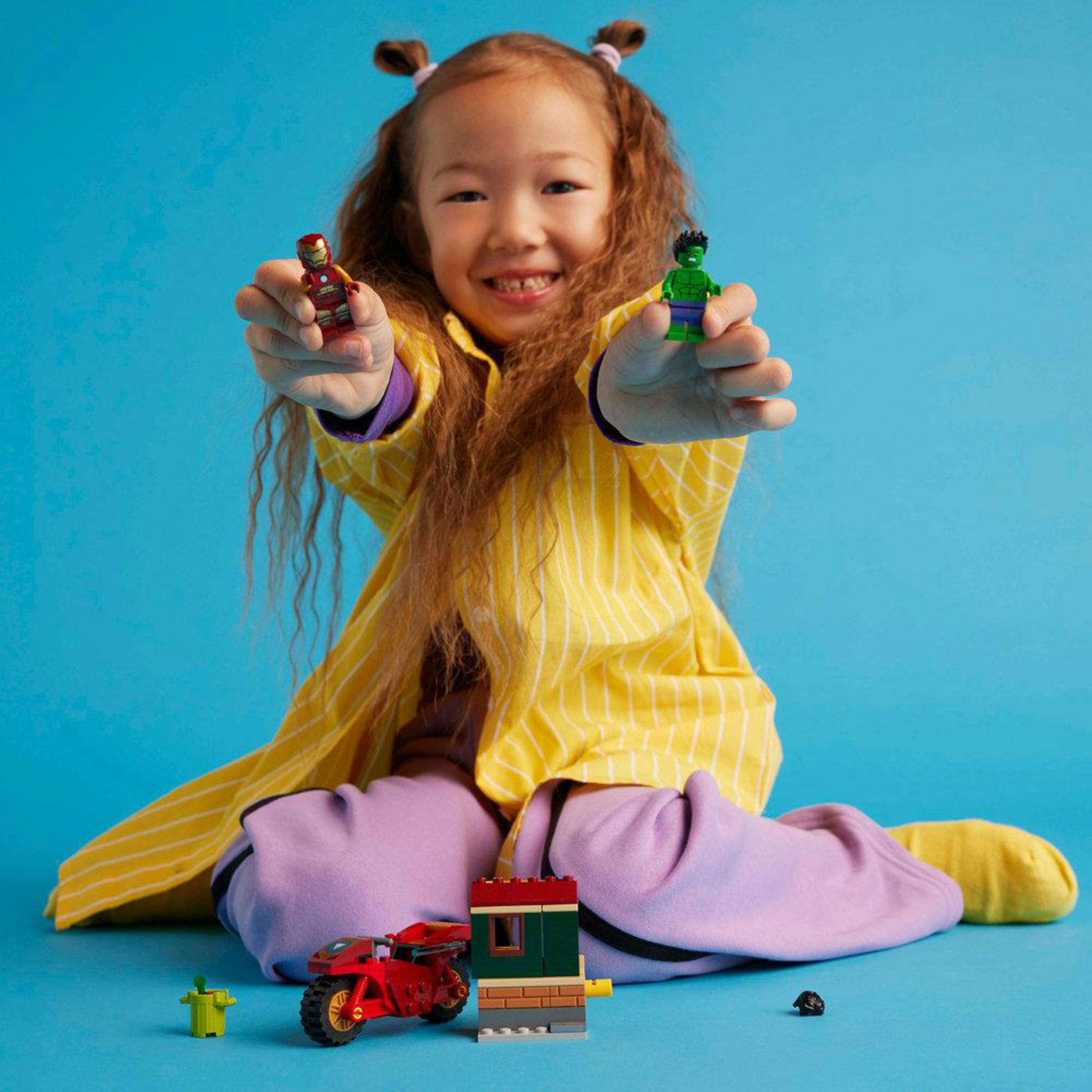Dziewczyna trzymająca figurki Lego Iron Mana i Hulka, z zabawkami Lego na niebieskim tle.