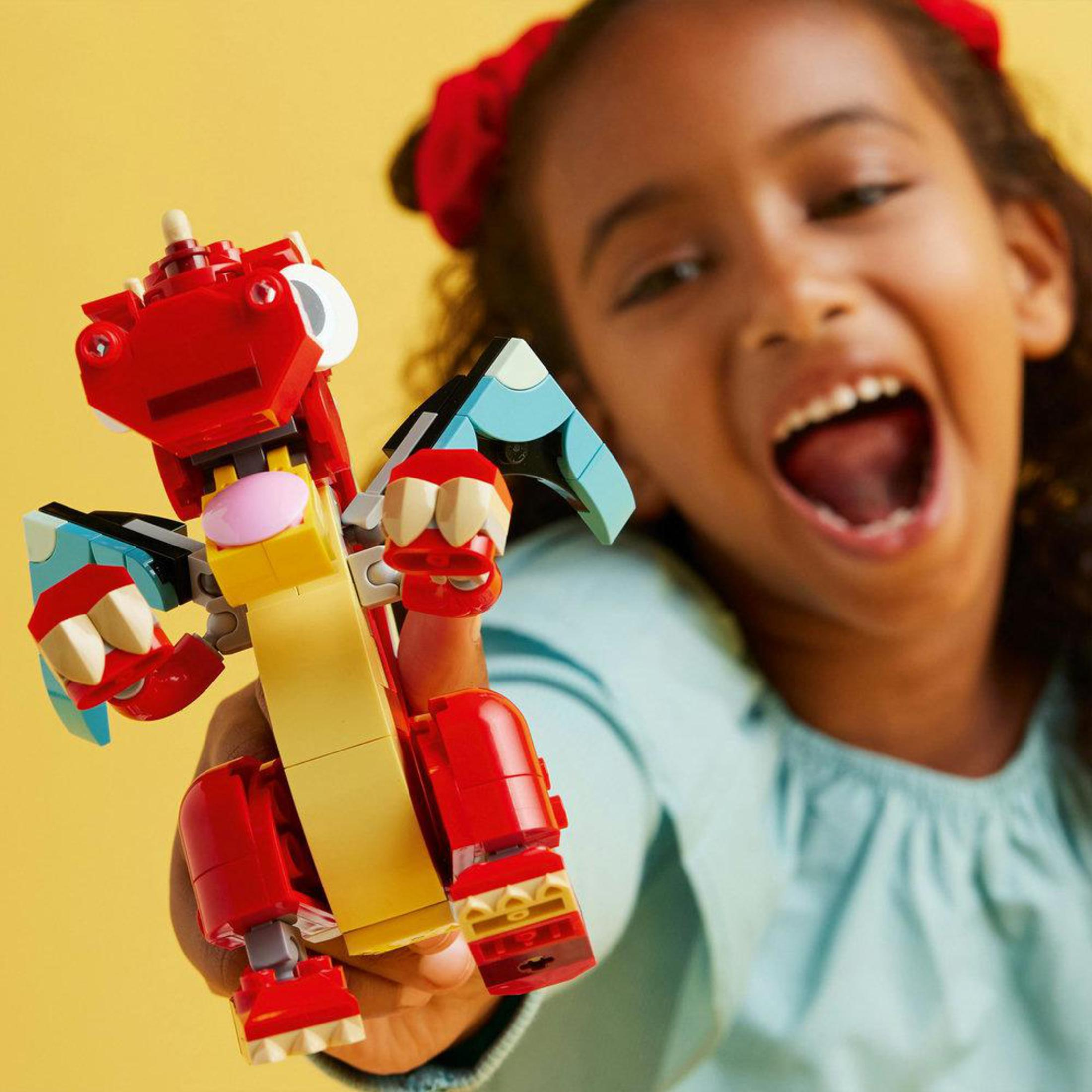Una niña sosteniendo un dragón Lego rojo, sonriendo ampliamente. Fondo amarillo.