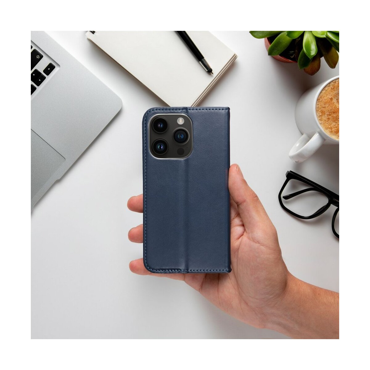 Mano che tiene custodia blu navy con laptop, taccuino, penna, pianta e tazza.