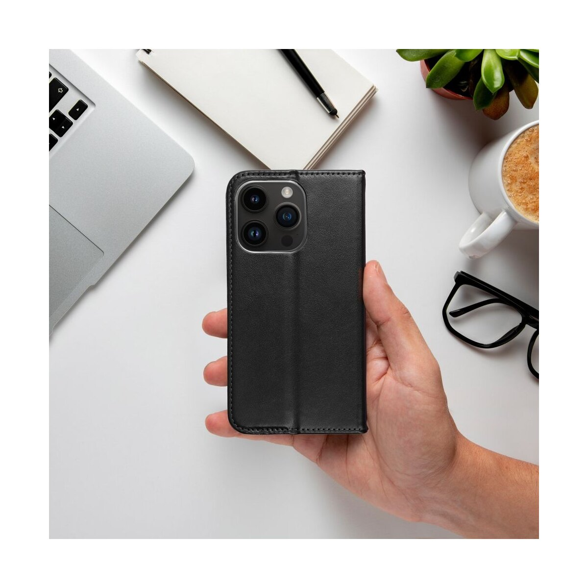 Una persona tiene in mano una custodia nera con un telefono dentro, accanto a un laptop, una tazza di caffè e occhiali.