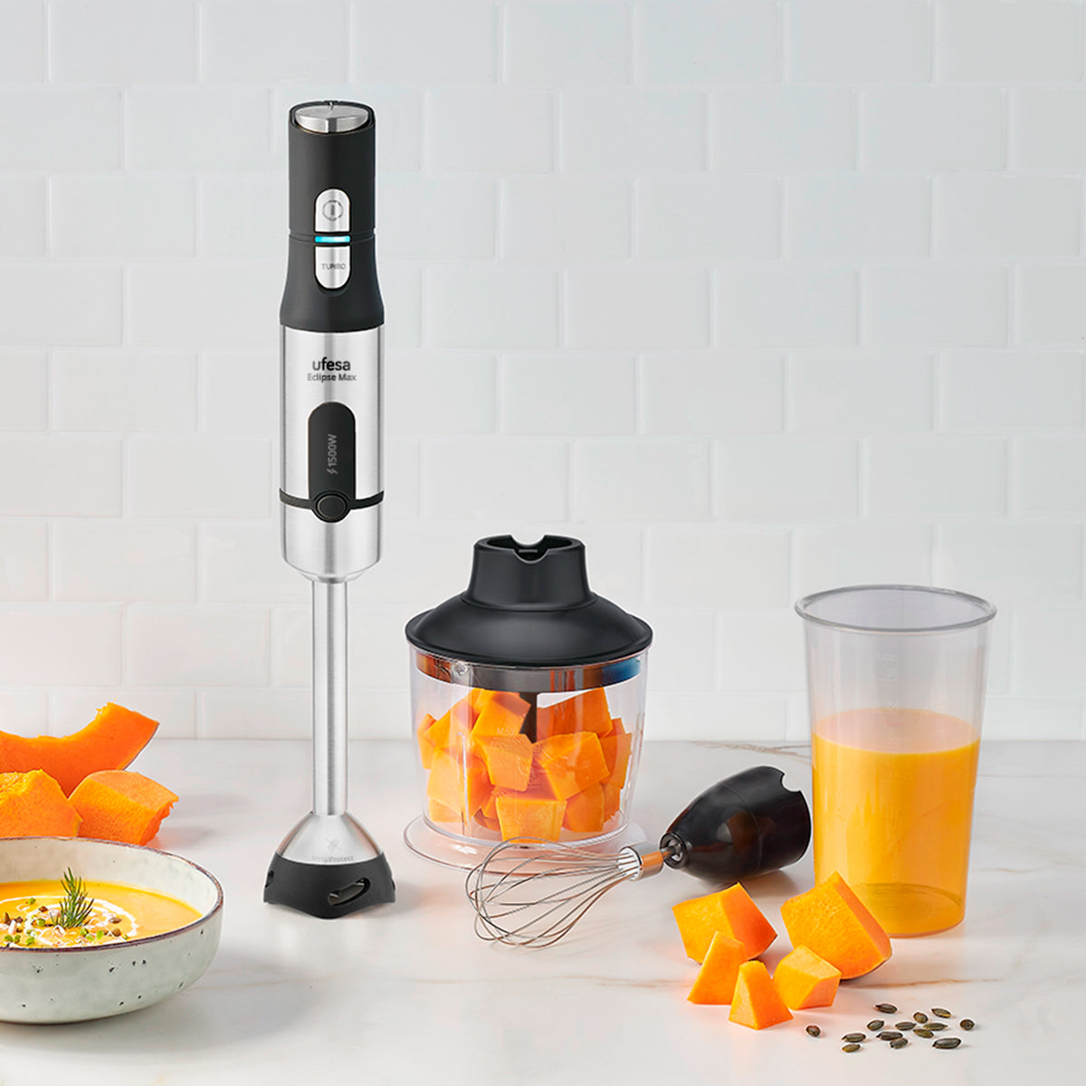 Un frullatore a immersione con zucca arancione e succo in un bicchiere su superficie piastrellata bianca.
