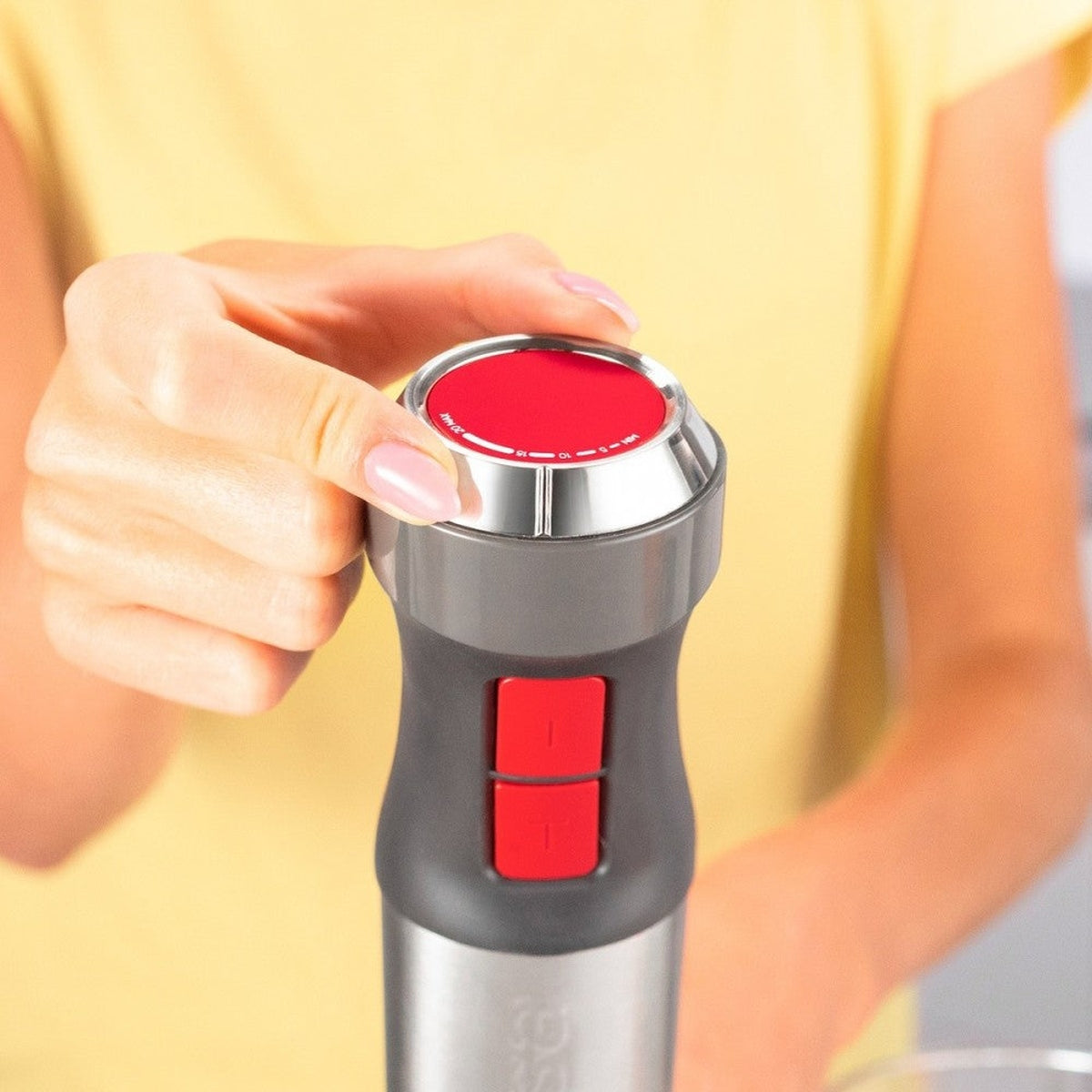 Ręka reguluje czerwone pokrętło na szaro-czarnym blenderze ręcznym, żółte tło.