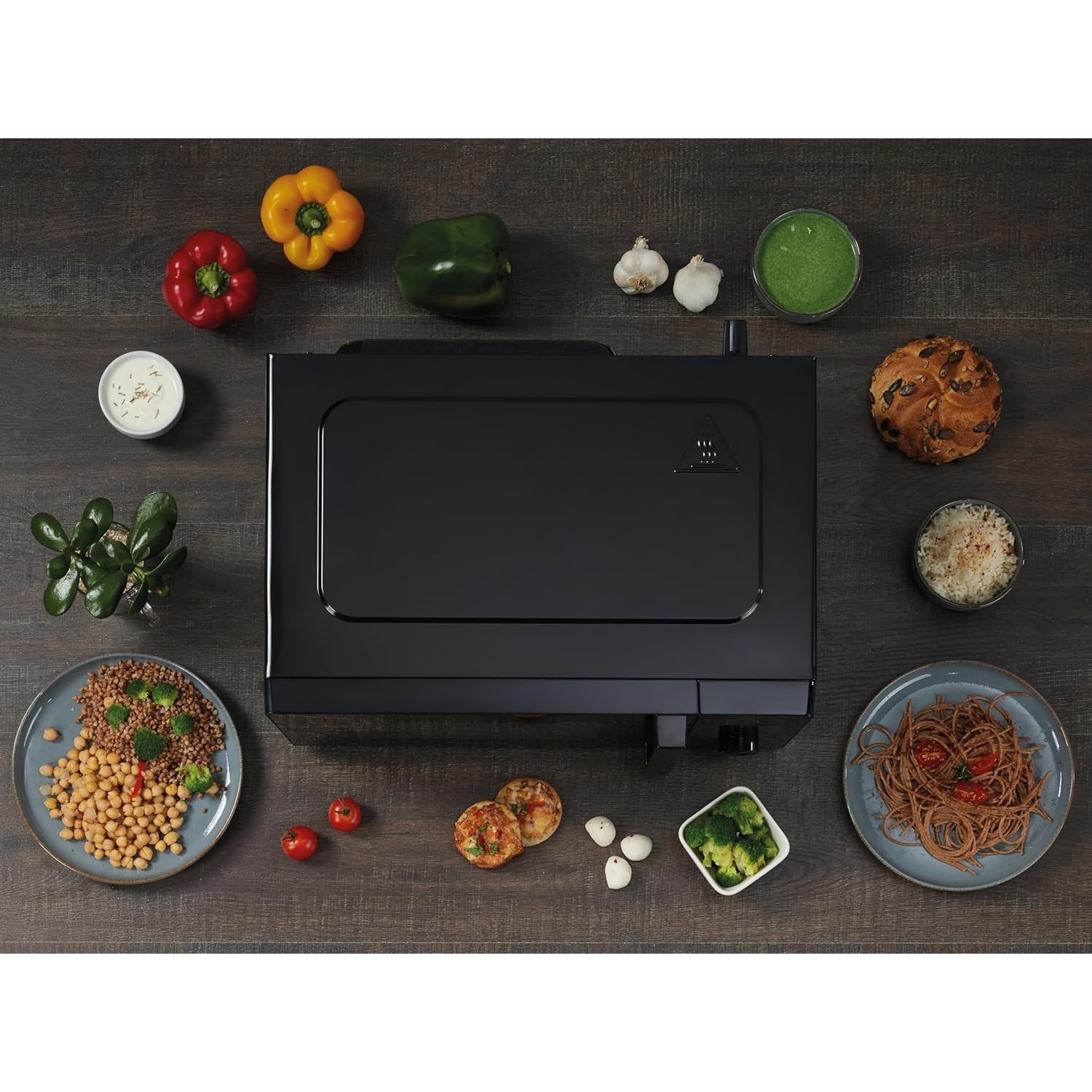 Vista cenital de un electrodoméstico negro rodeado de alimentos: pimientos, verduras y platos.