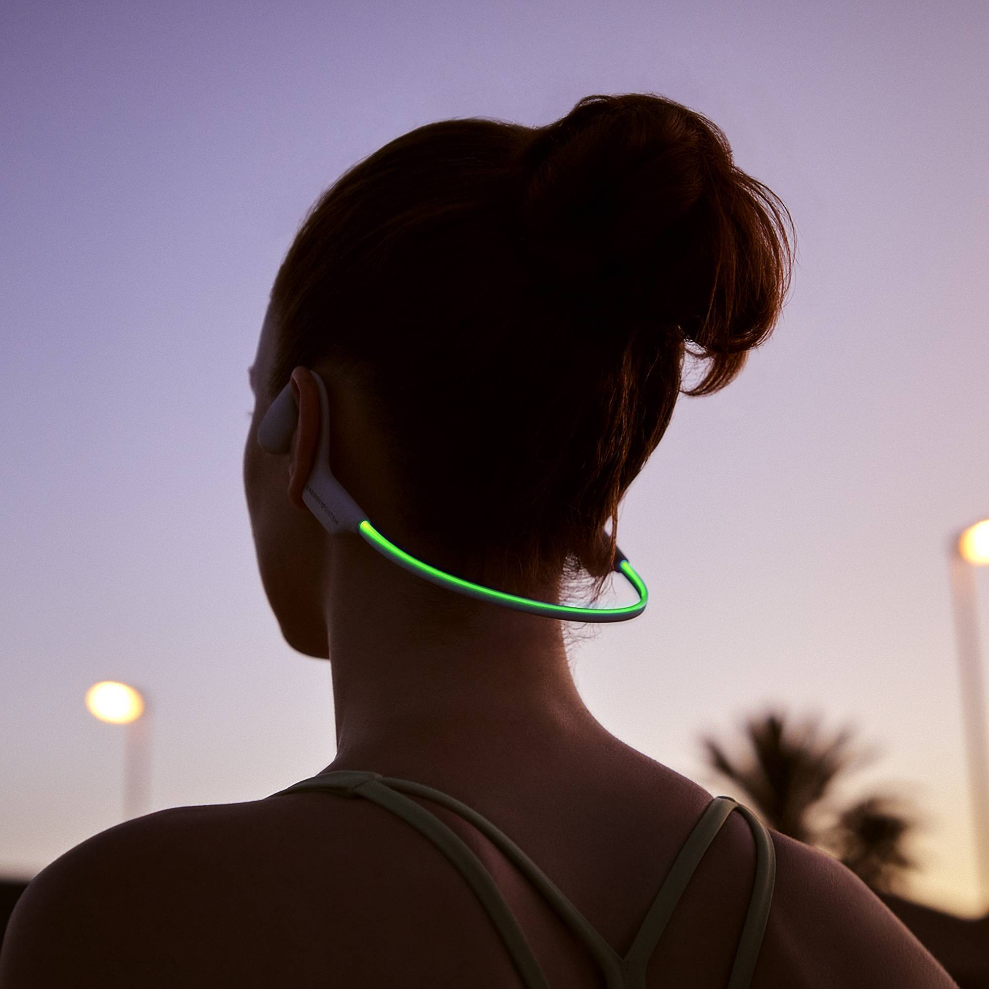 Mujer con auriculares, silueta contra el atardecer. Luz verde alrededor del cuello.