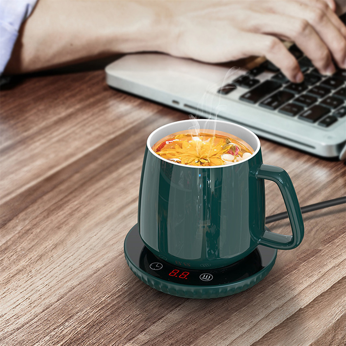 Grüner Becher auf einem Wärmer, mit Dampf. Laptop und Hände im Hintergrund.
