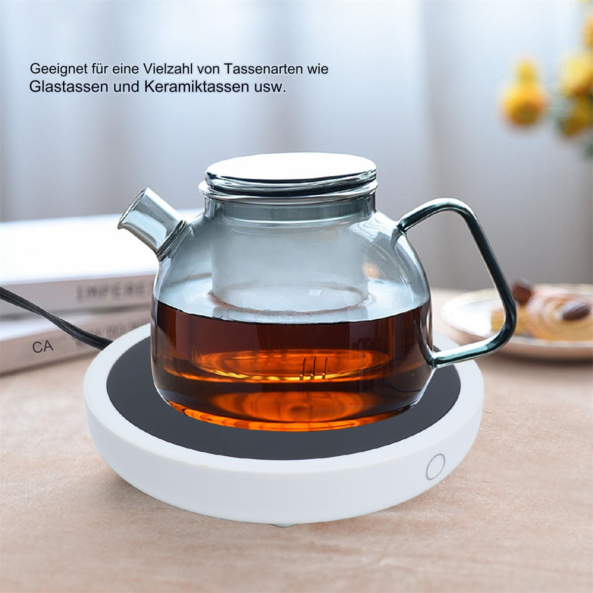 Gläserne Teekanne auf weißer Warmhaltebasis mit schwarzem Kabel, auf einem Tisch.