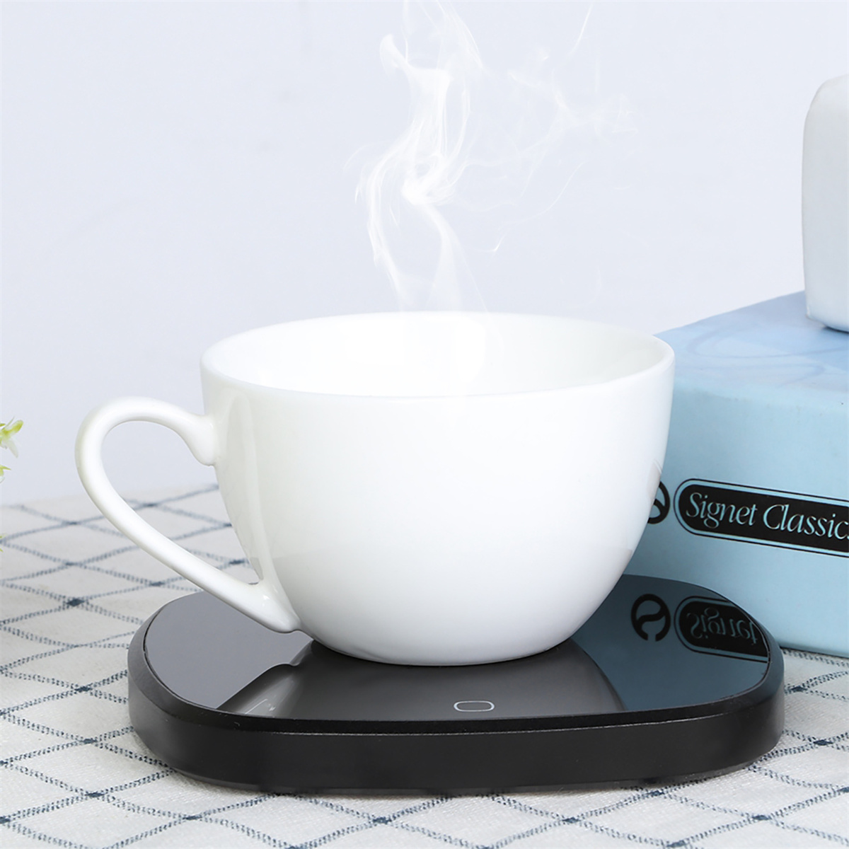 Weiße Tasse auf schwarzem Wärmer, Dampf steigt auf, im Hintergrund eine hellblaue Box.
