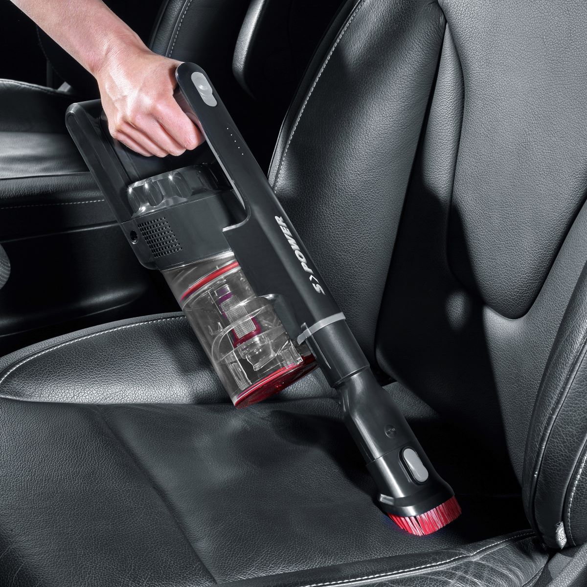 Una persona usando una aspiradora inalámbrica negra y roja en un asiento de cuero negro.