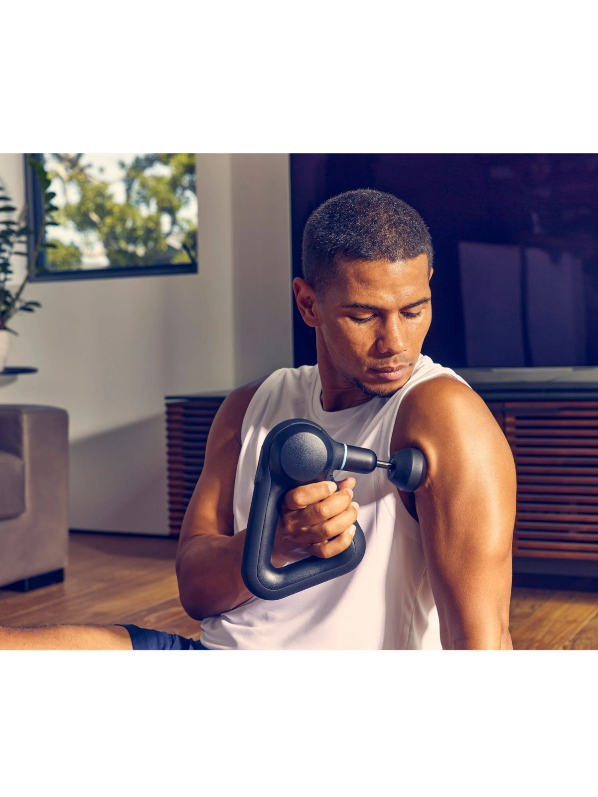 Mężczyzna używa czarnego pistoletu do masażu na ramieniu. TV i sofa w tle.