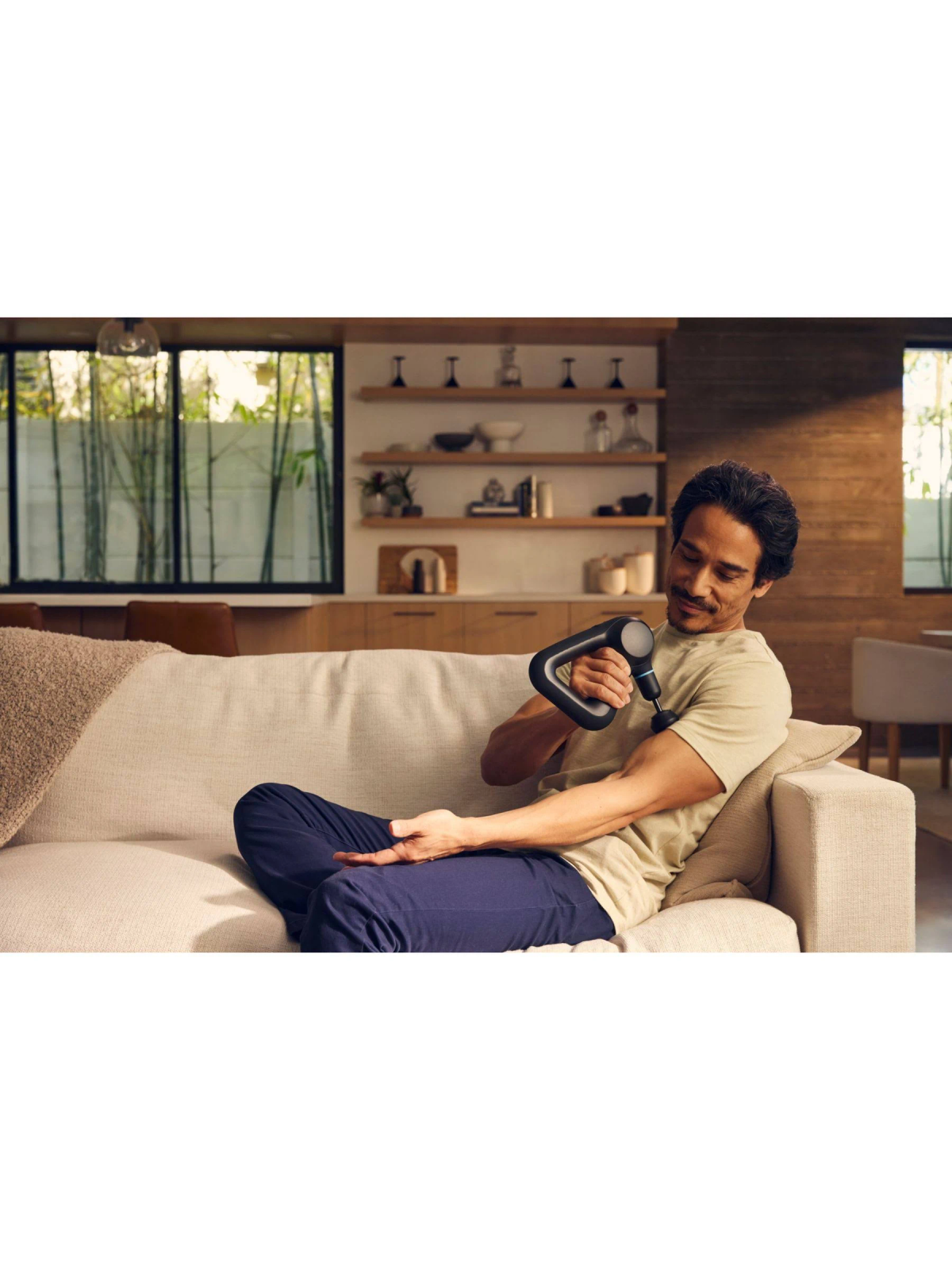 Mężczyzna używający czarnego pistoletu do masażu na ramieniu, siedząc na kanapie.