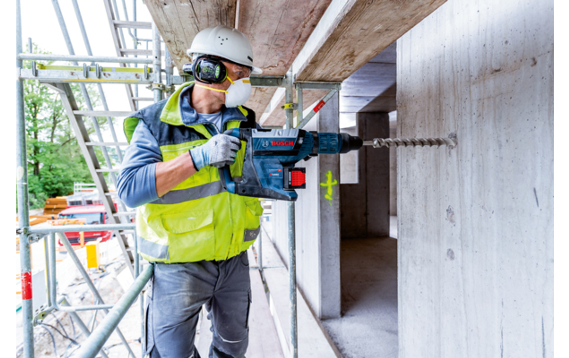 Trabajador de la construcción taladra hormigón con martillo Bosch. Andamios al fondo.