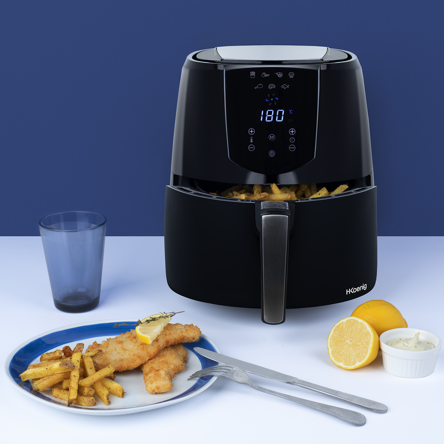Una friggitrice ad aria nera con cibo all'interno, più altri alimenti su un tavolo.