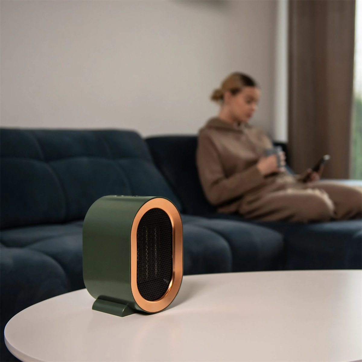Grüner und bronzener elektrischer Heizlüfter auf einem weißen Tisch, mit einer Frau auf einem blauen Sofa im Hintergrund.