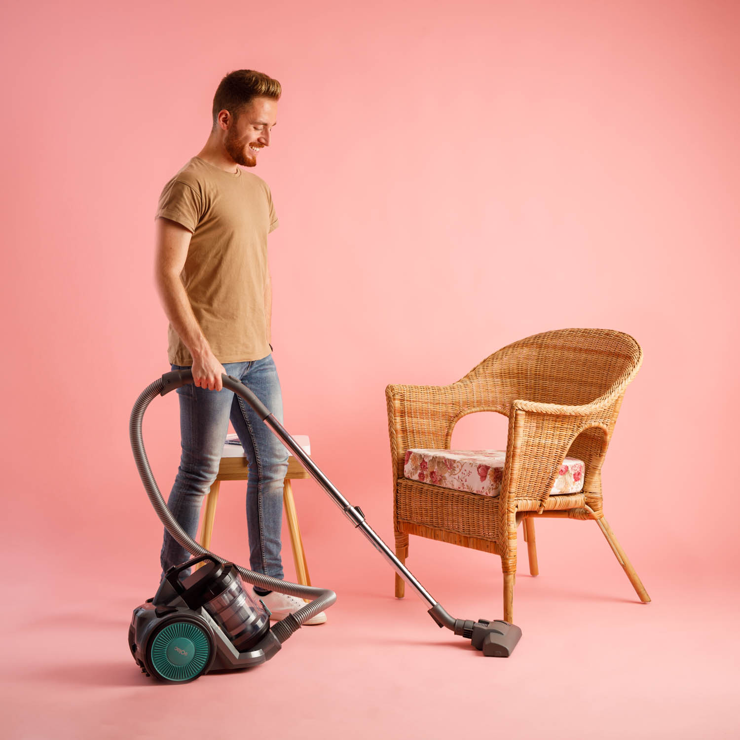 Mężczyzna odkurza pokój. Ma na sobie casualowe ubrania. Widoczny jest wiklinowy fotel i różowe tło.