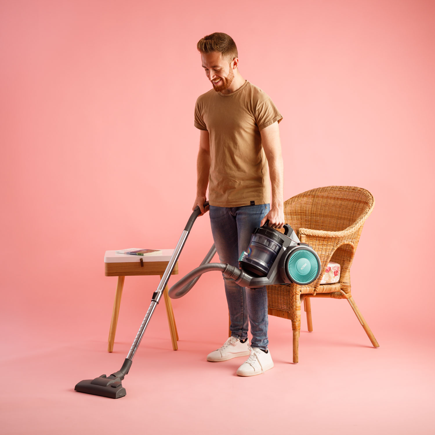 Mężczyzna odkurza pokój z różowym tłem. Trzyma odkurzacz. Jest krzesło i stolik.