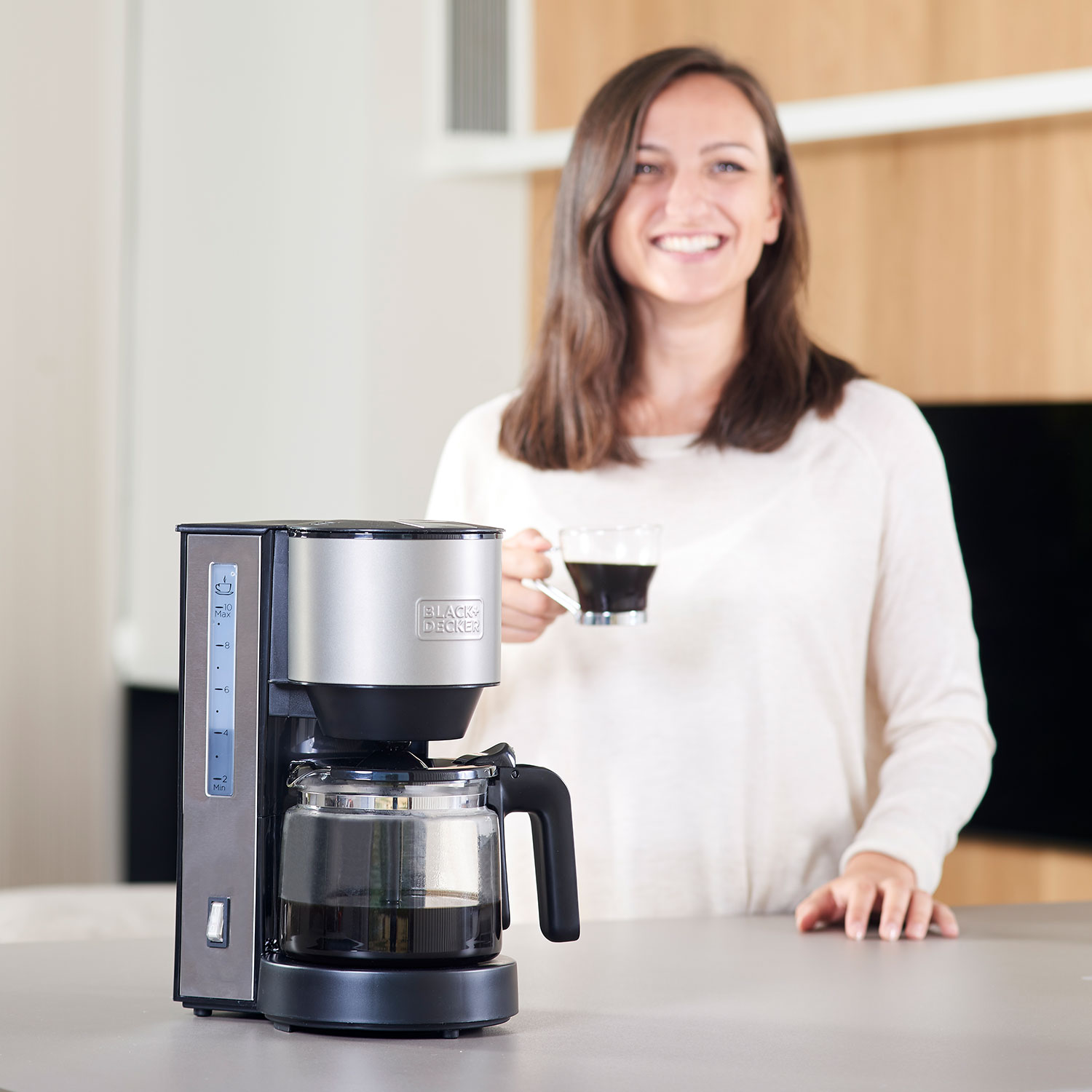 Kobieta uśmiecha się, trzymając filiżankę kawy w pobliżu ekspresu do kawy.