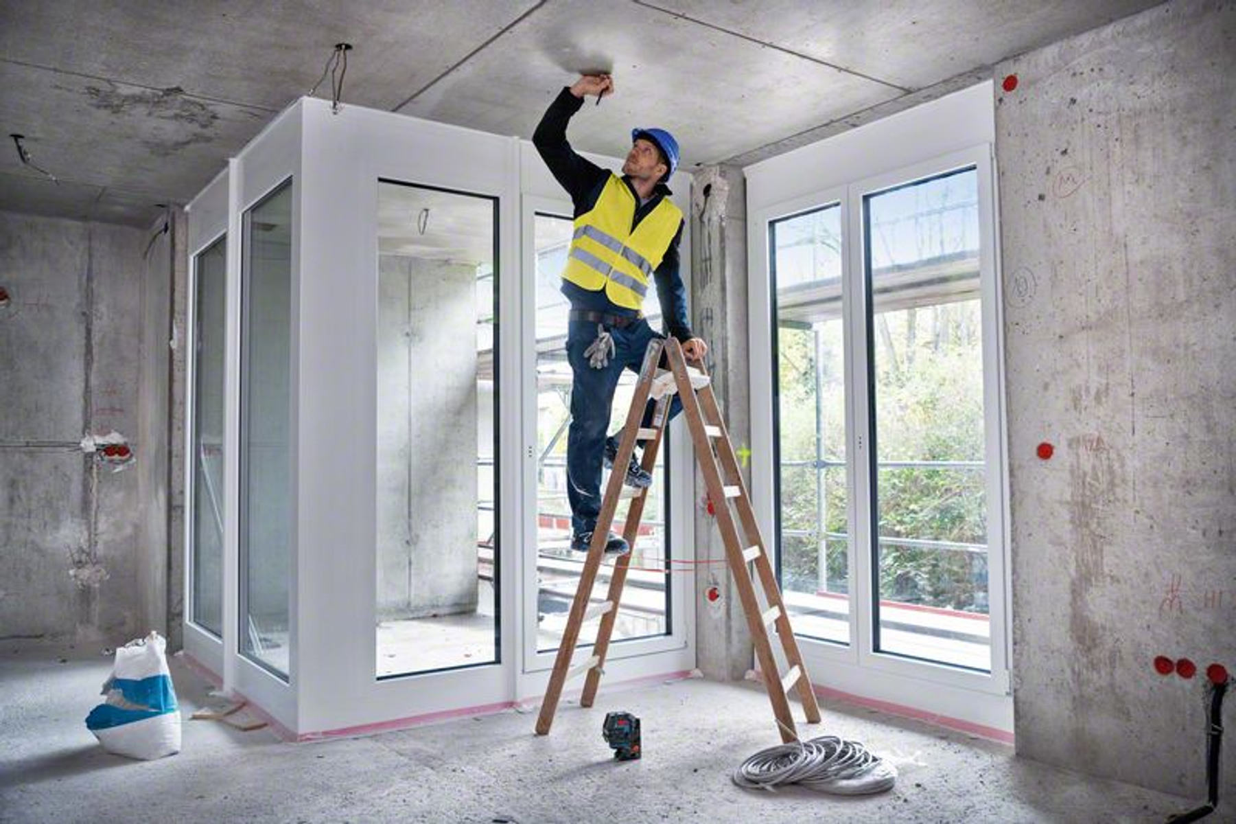 Wnętrze placu budowy z mężczyzną na drabinie. Mężczyzna pracuje nad sufitem, ma na sobie kamizelkę i kask.