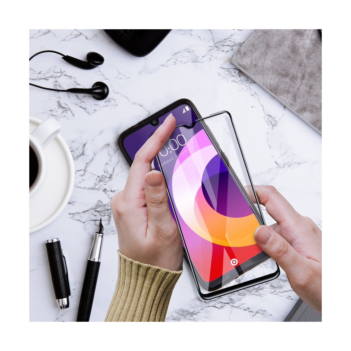 Mani che tengono un telefono con una protezione schermo applicata. Altri elementi: penna, cuffie, tazza.