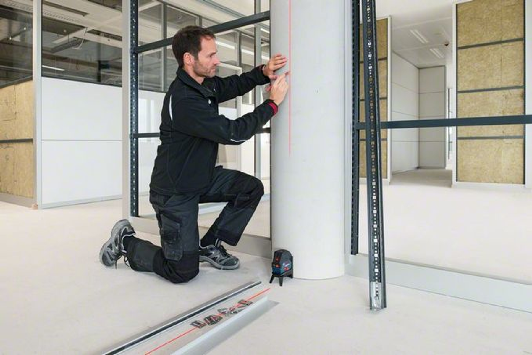 Mężczyzna klęczy, używając poziomicy do zaznaczenia czerwonej linii na białej kolumnie. Miejsce budowy.