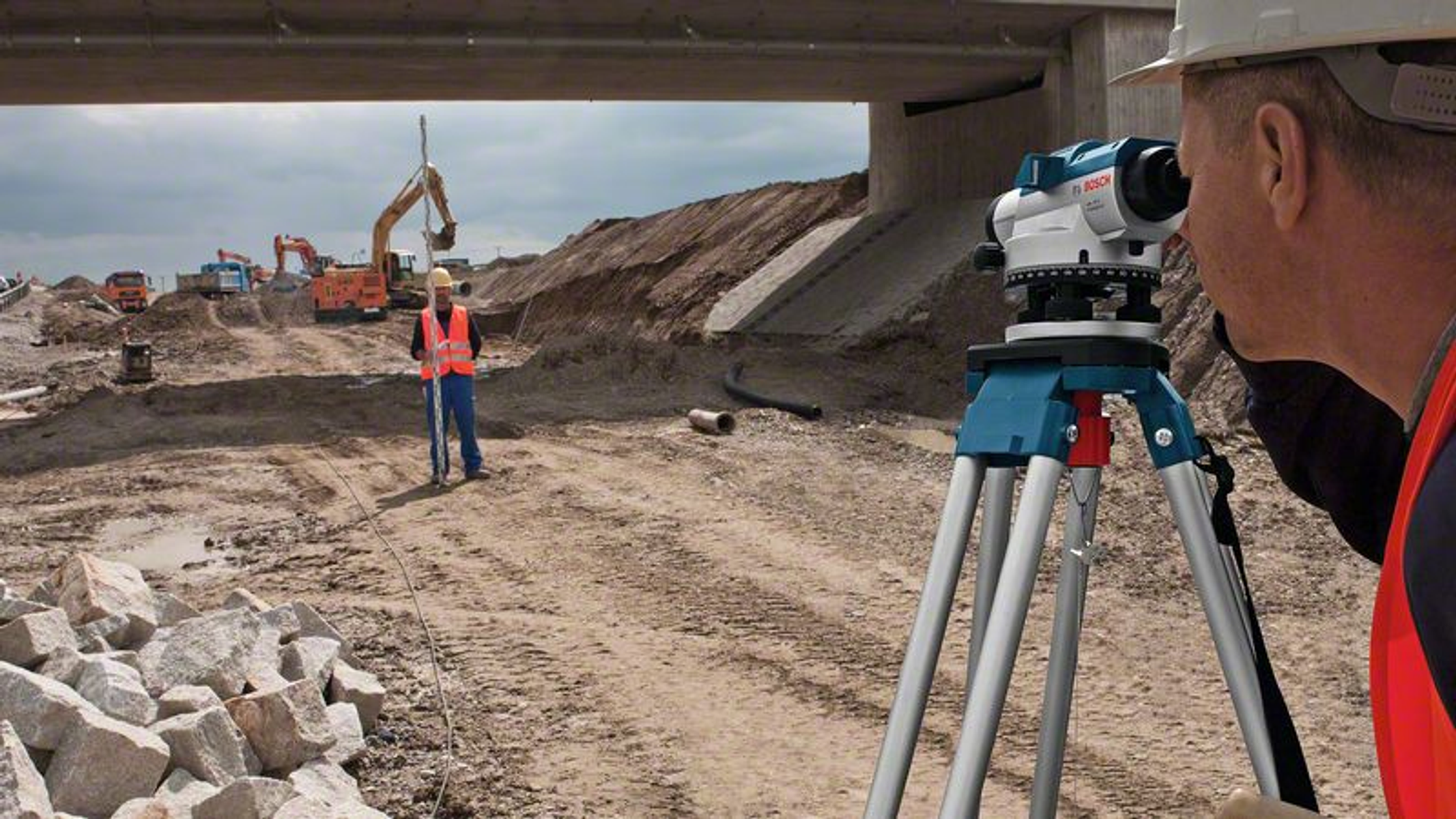 Geodeta patrzy przez teodolit. Plac budowy z maszynami i pod mostem w tle.