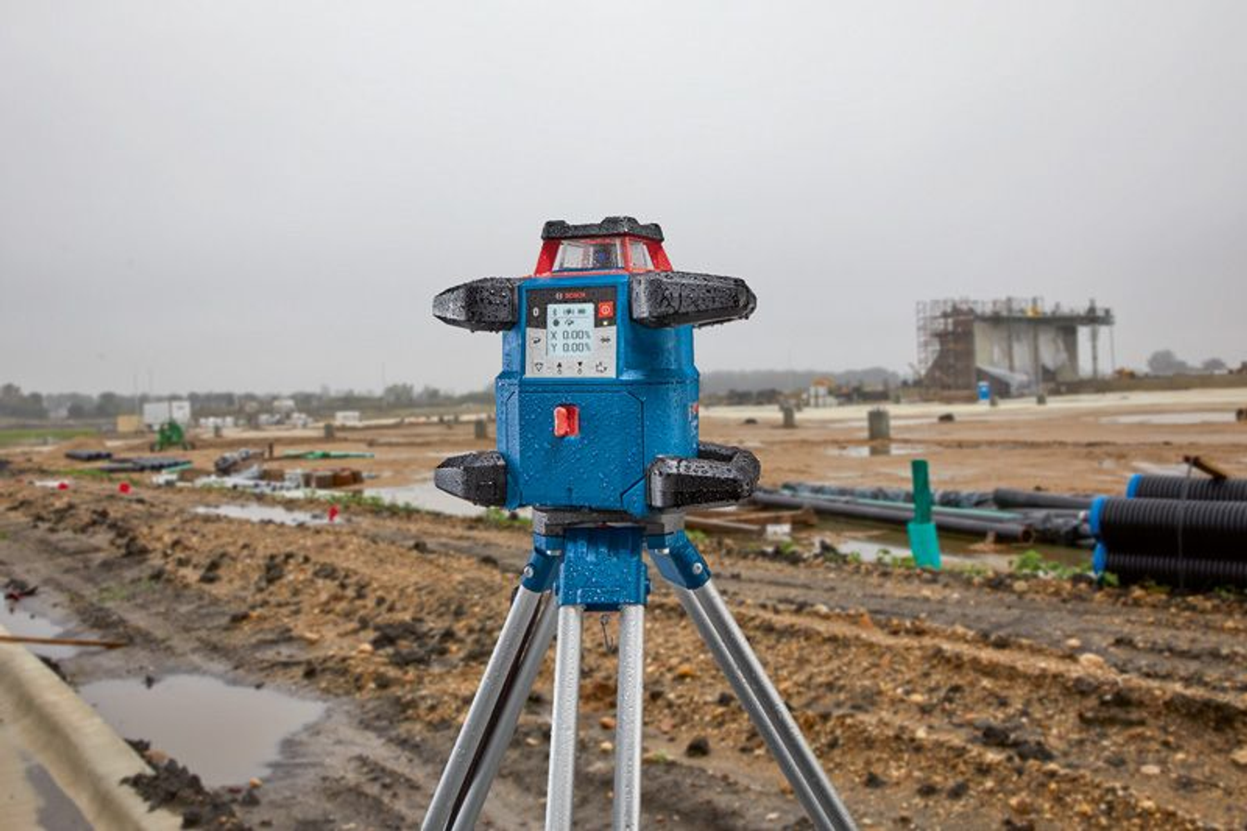 Un nivel láser azul y plateado sobre un trípode, en una obra en construcción con cielos nublados.