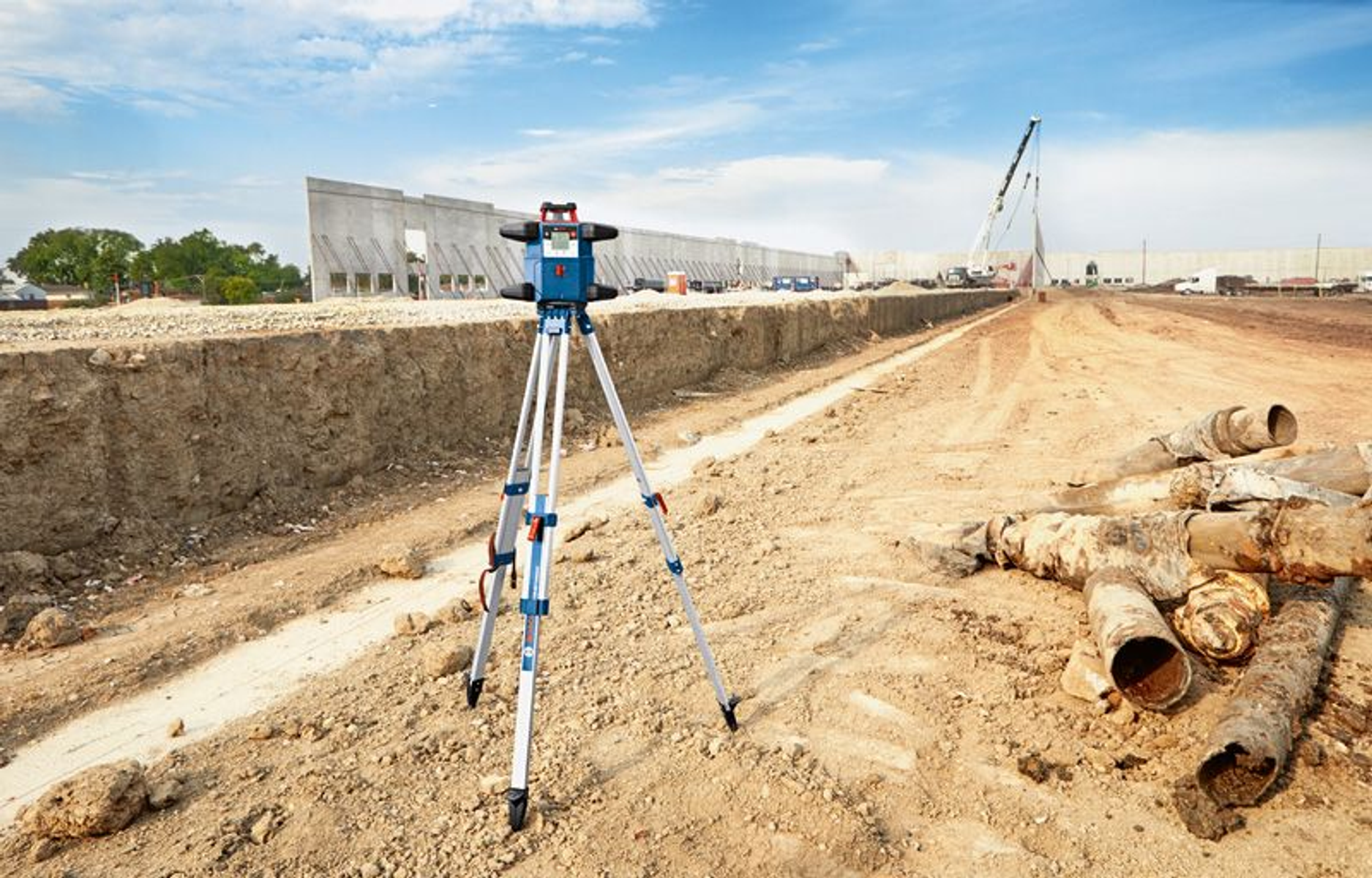 Sitio de construcción con un nivel láser en un trípode. Una grúa y edificios al fondo.