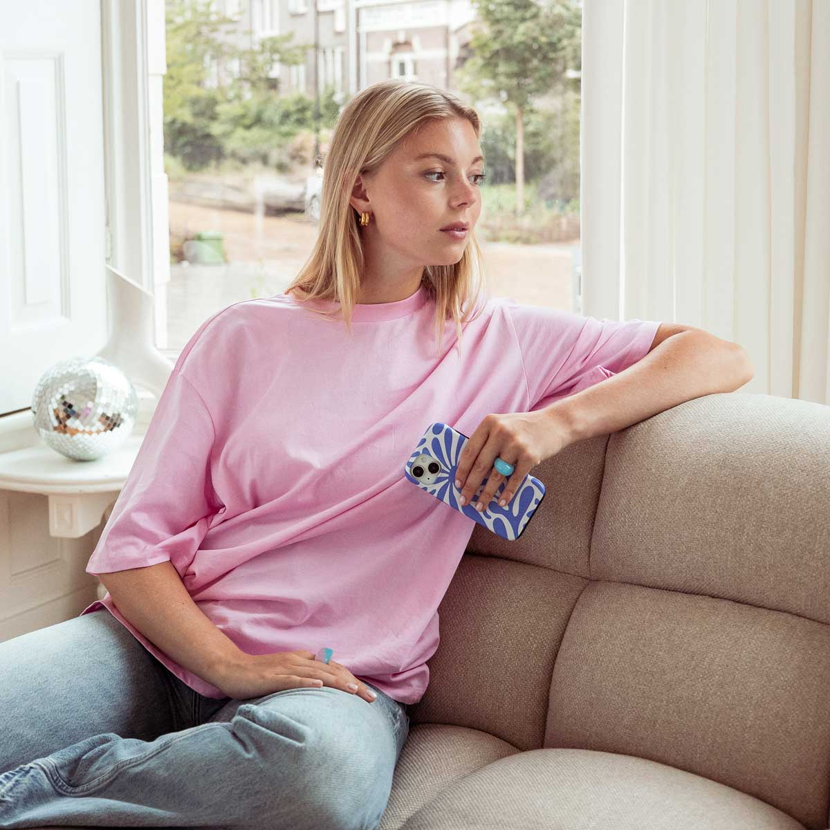 Femme assise sur un canapé beige, tenant un téléphone avec une coque bleue et blanche. Chemise rose, vue fenêtre.