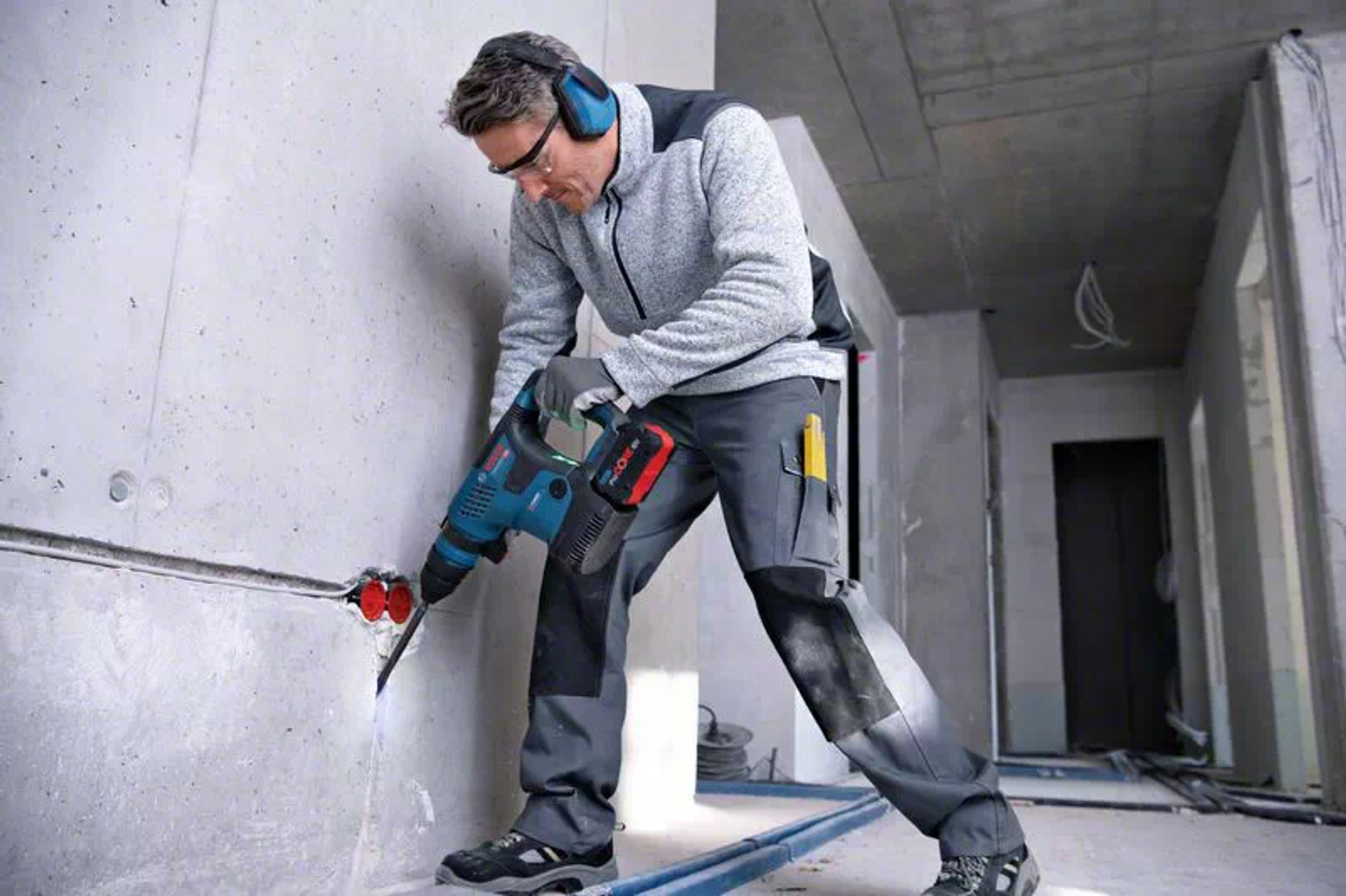 Osoba wiercąca w ścianie niebiesko-czarną wiertarką Bosch, w odzieży ochronnej.