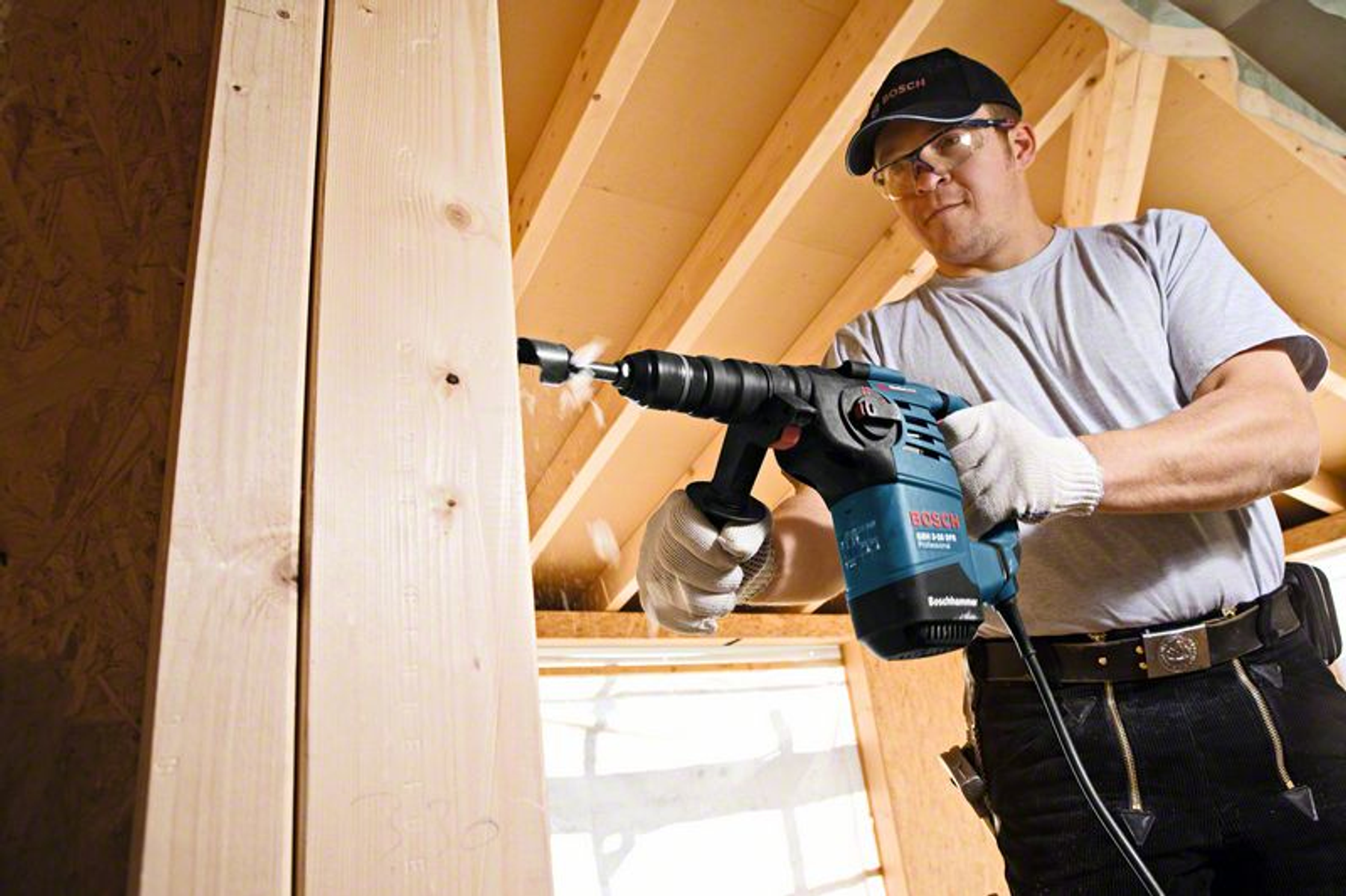 Mężczyzna wiercący w drewnie wiertarką Bosch, w rękawicach i czapce, w pomieszczeniu.