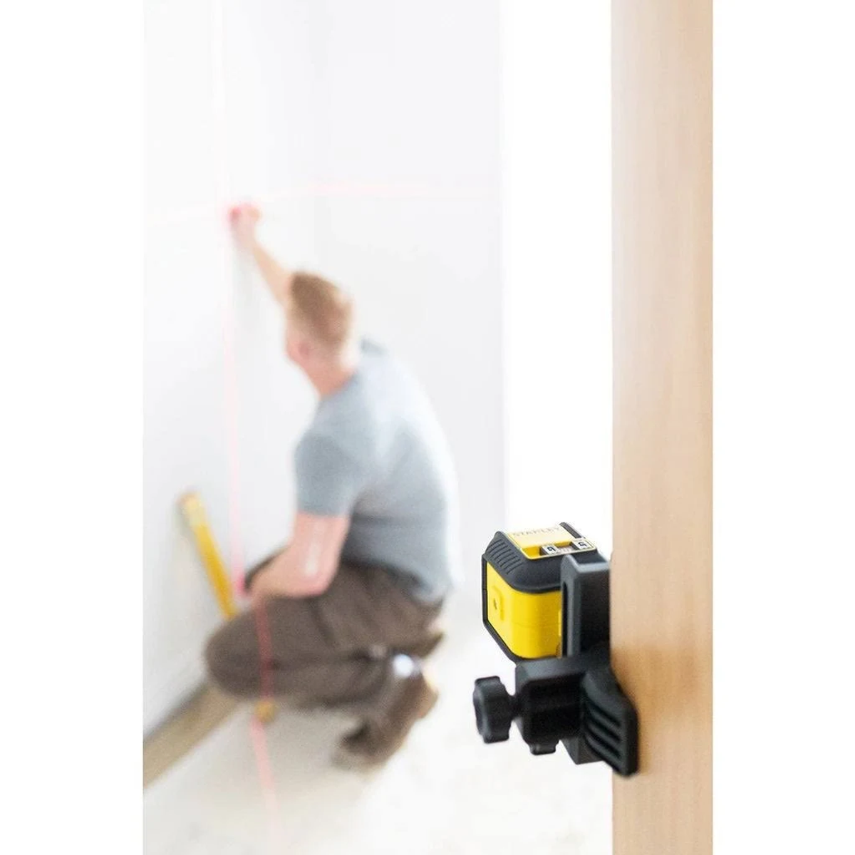 Żółto-czarna poziomica laserowa przymocowana do drewnianej ramy drzwi. Osoba pracuje w tle.