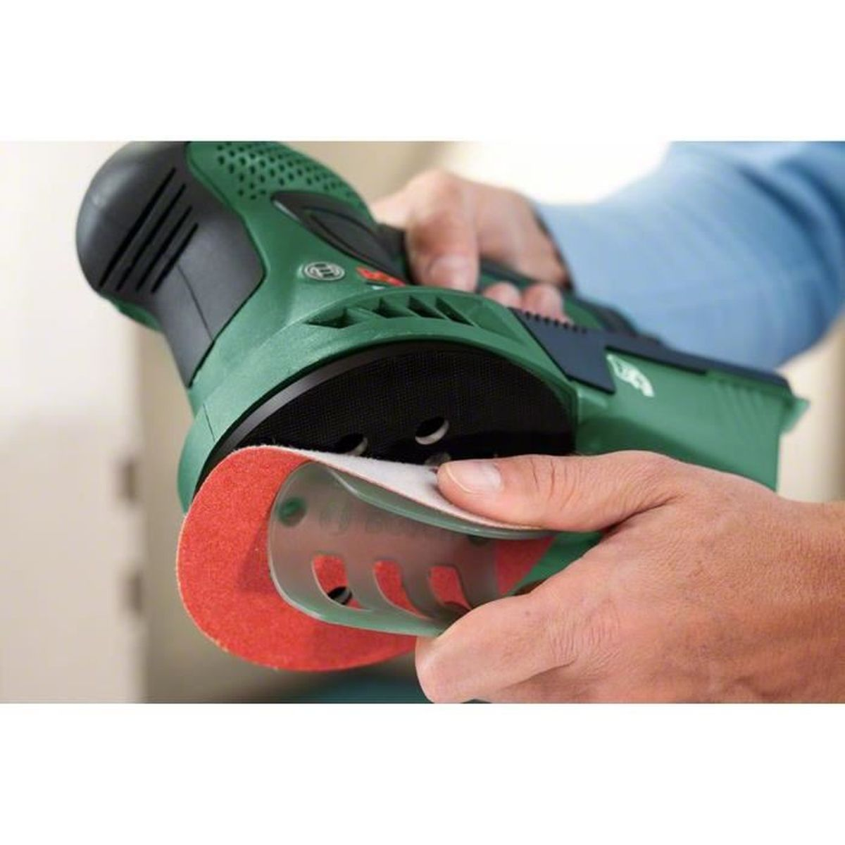 Mains d'une personne tenant une ponceuse verte avec un disque de ponçage rouge. L'outil est en cours d'utilisation.