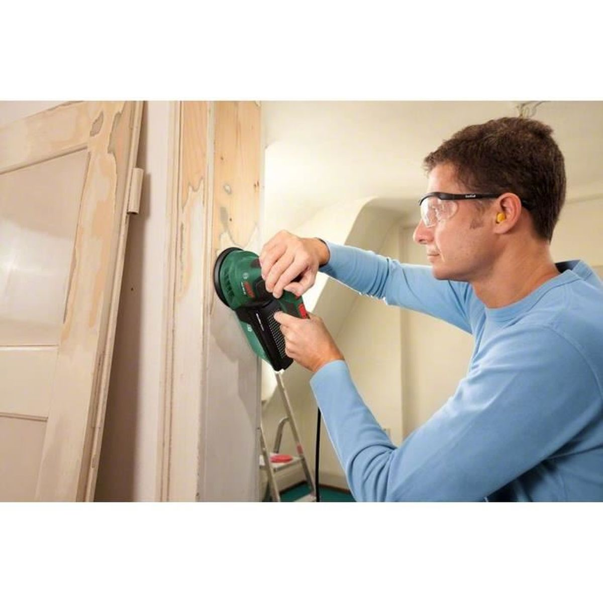 Homme ponçant une porte avec une ponceuse Bosch, portant lunettes et bouchons d'oreille.