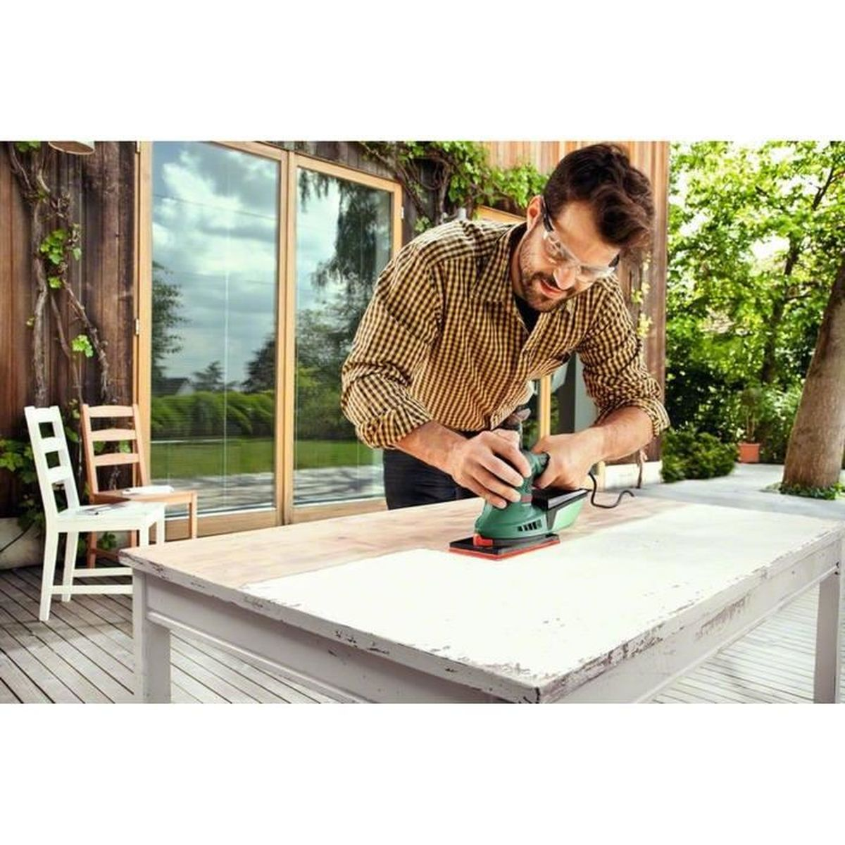 Un homme ponce une table en bois à l'extérieur avec une ponceuse. Une chaise et du feuillage sont en arrière-plan.