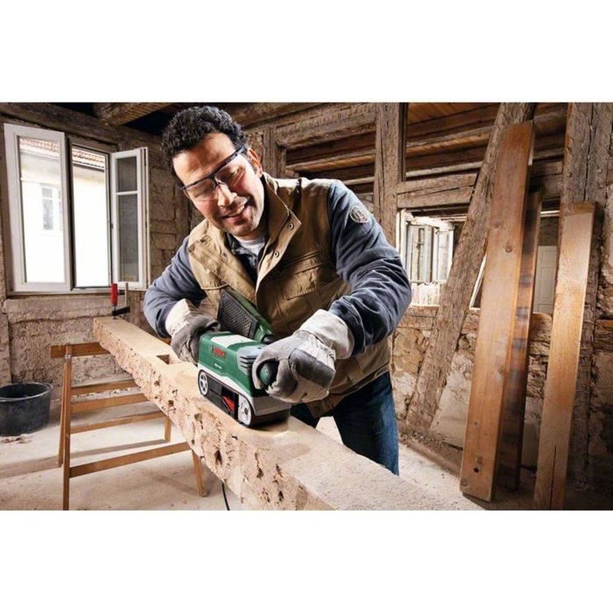 Un homme utilisant une ponceuse sur une poutre en bois dans un cadre rustique.