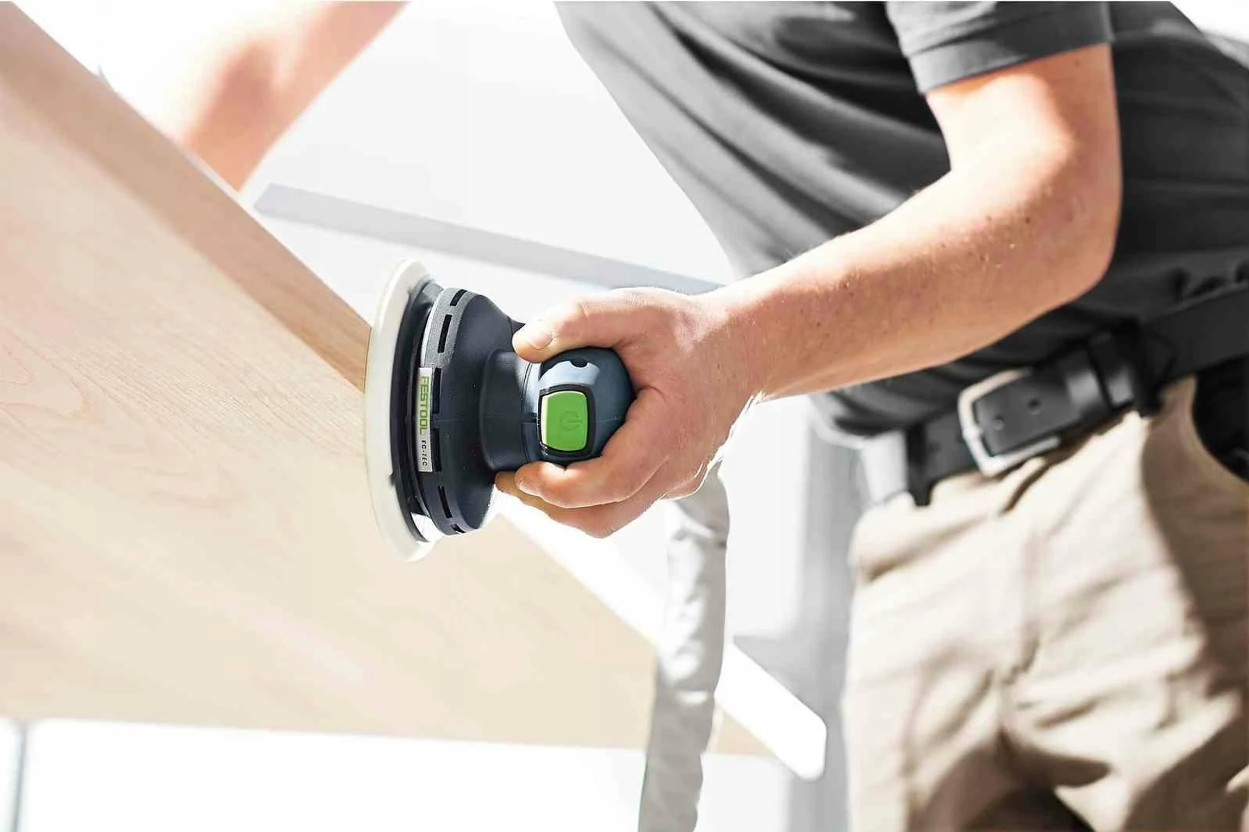 Persona usando una lijadora eléctrica en una superficie de madera. La lijadora es negra y verde.