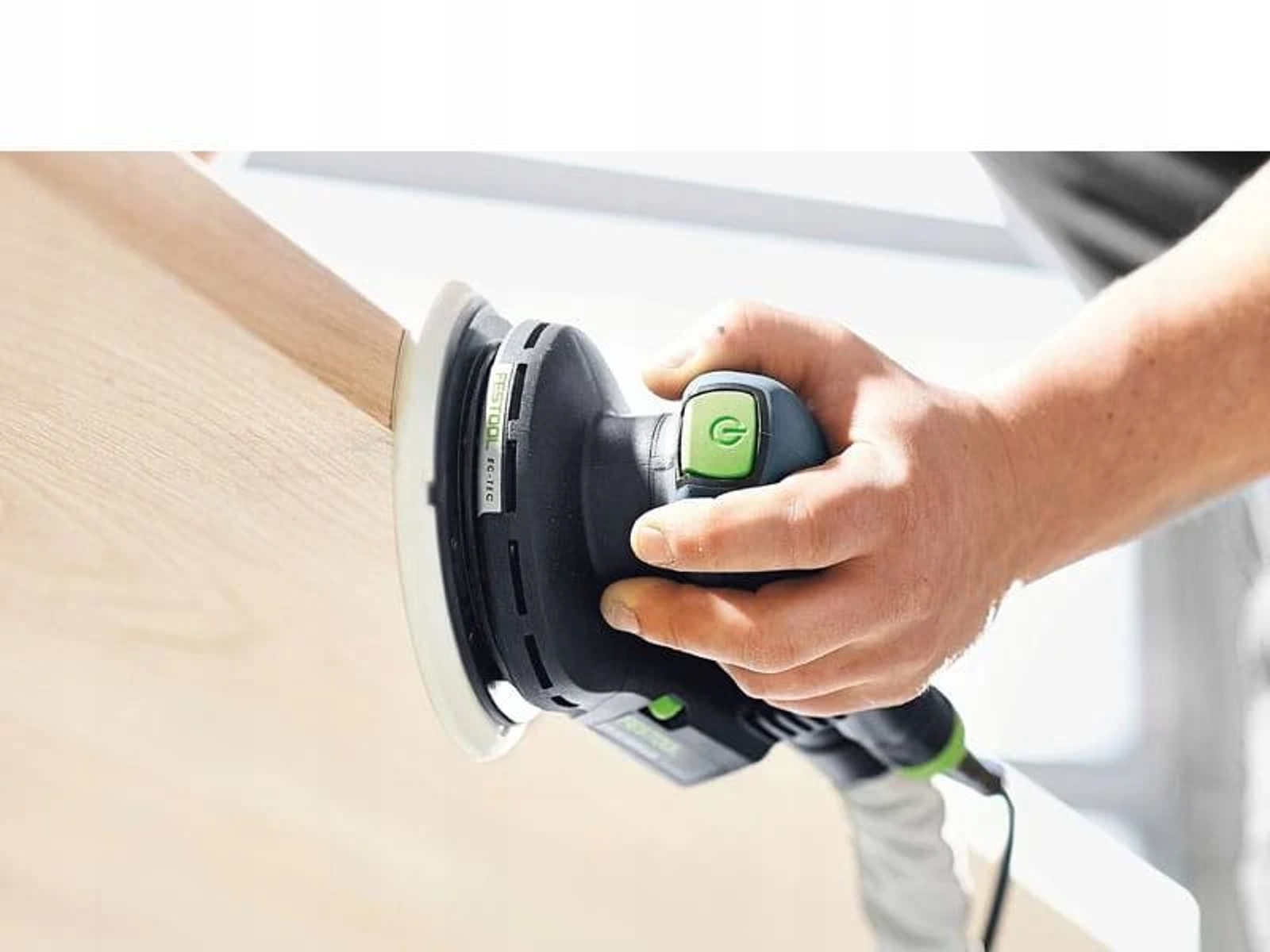 La mano de una persona sosteniendo una lijadora eléctrica negra y verde, lijando madera. La lijadora está cerca de la madera.