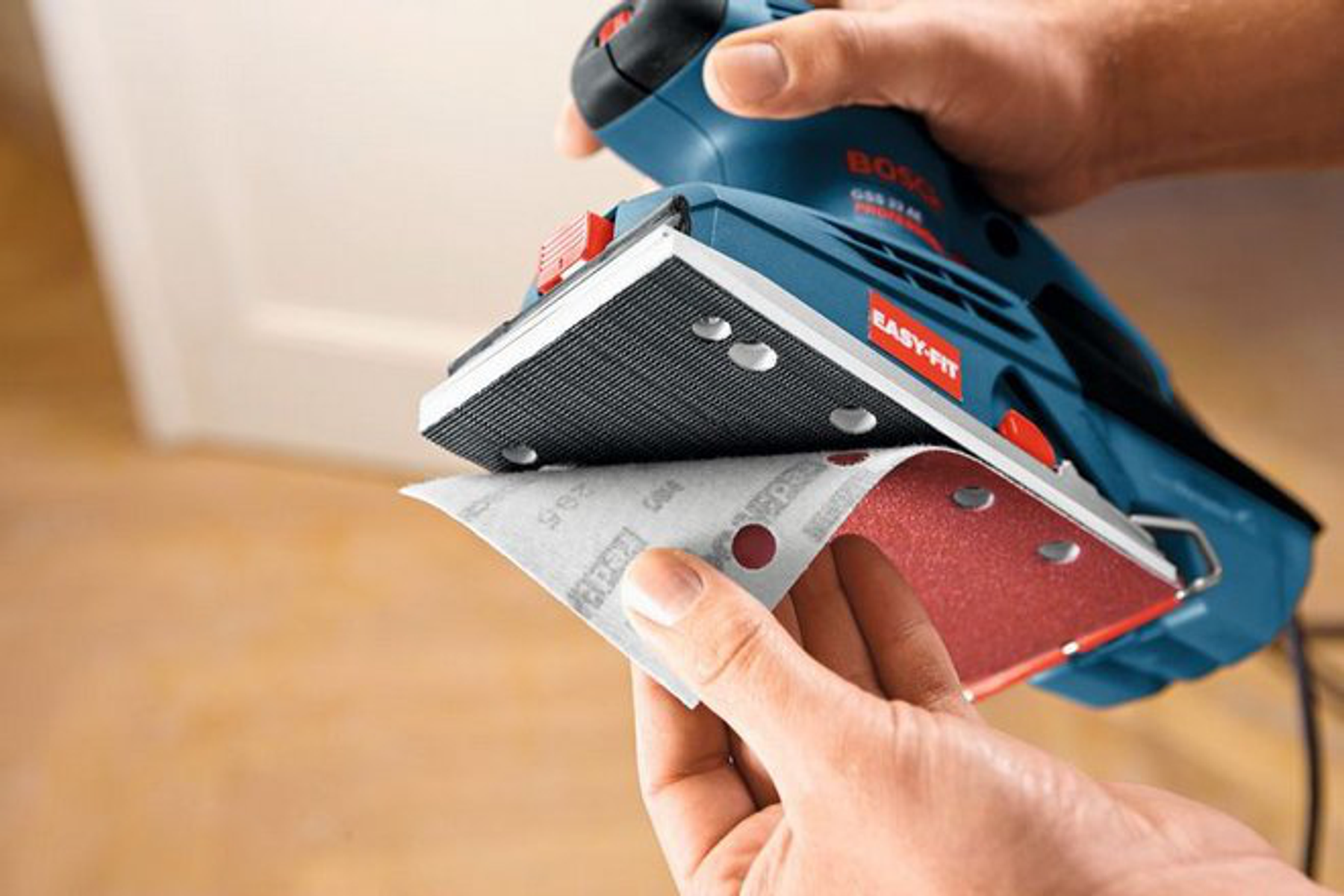 Mano sosteniendo una lijadora con papel de lija. Lijadora azul y roja, piso de madera al fondo.