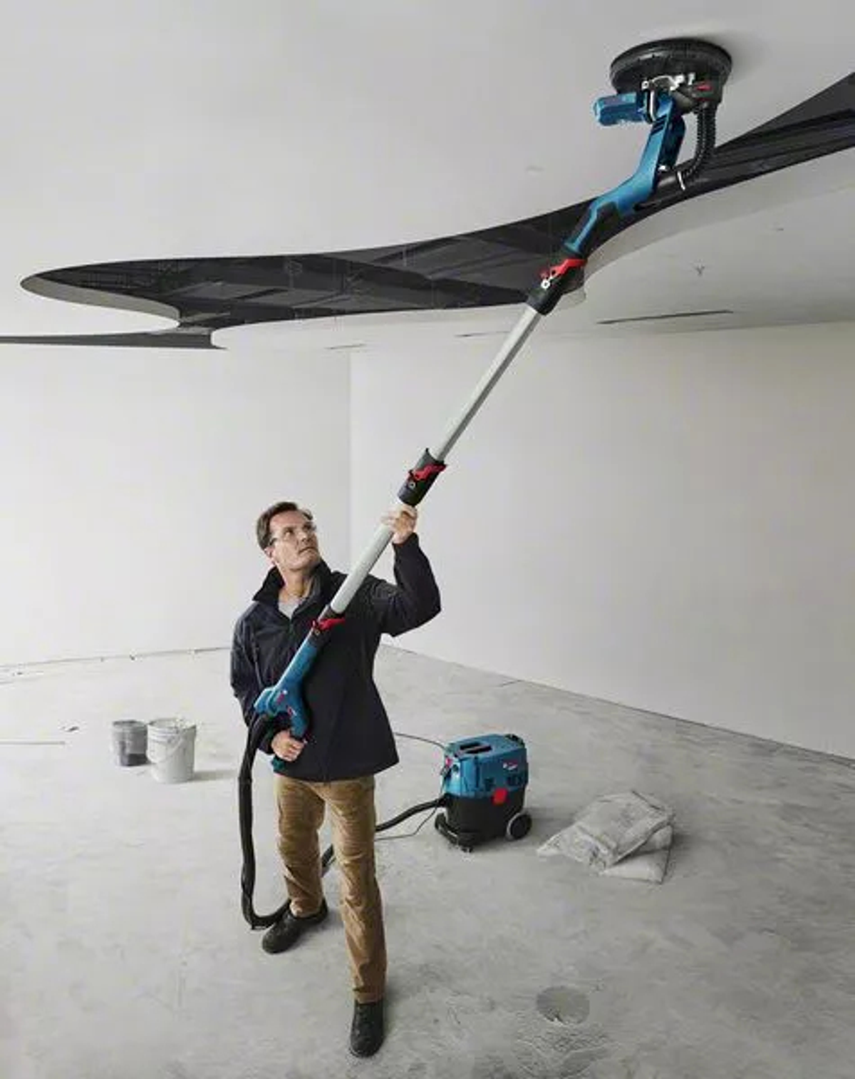 Homme ponçant un plafond avec une ponceuse à perche, murs blancs et aspirateur bleu.